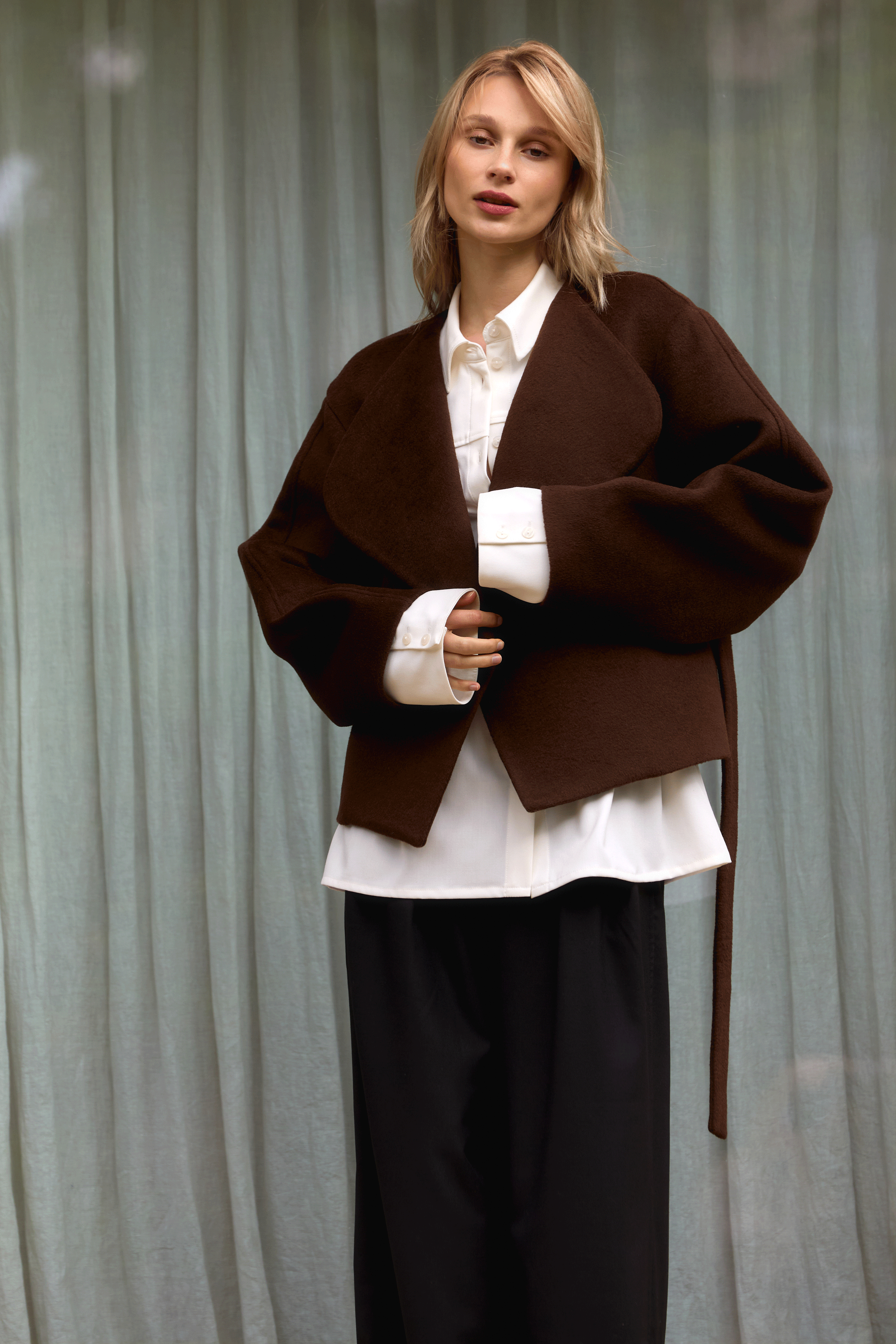 Model trägt eine braune, locker geschnittene Jacke mit breitem Revers und Bindegürtel über einer weißen Bluse mit Manschetten, kombiniert mit weiter schwarzer Hose, vor hellem Vorhang-Hintergrund.