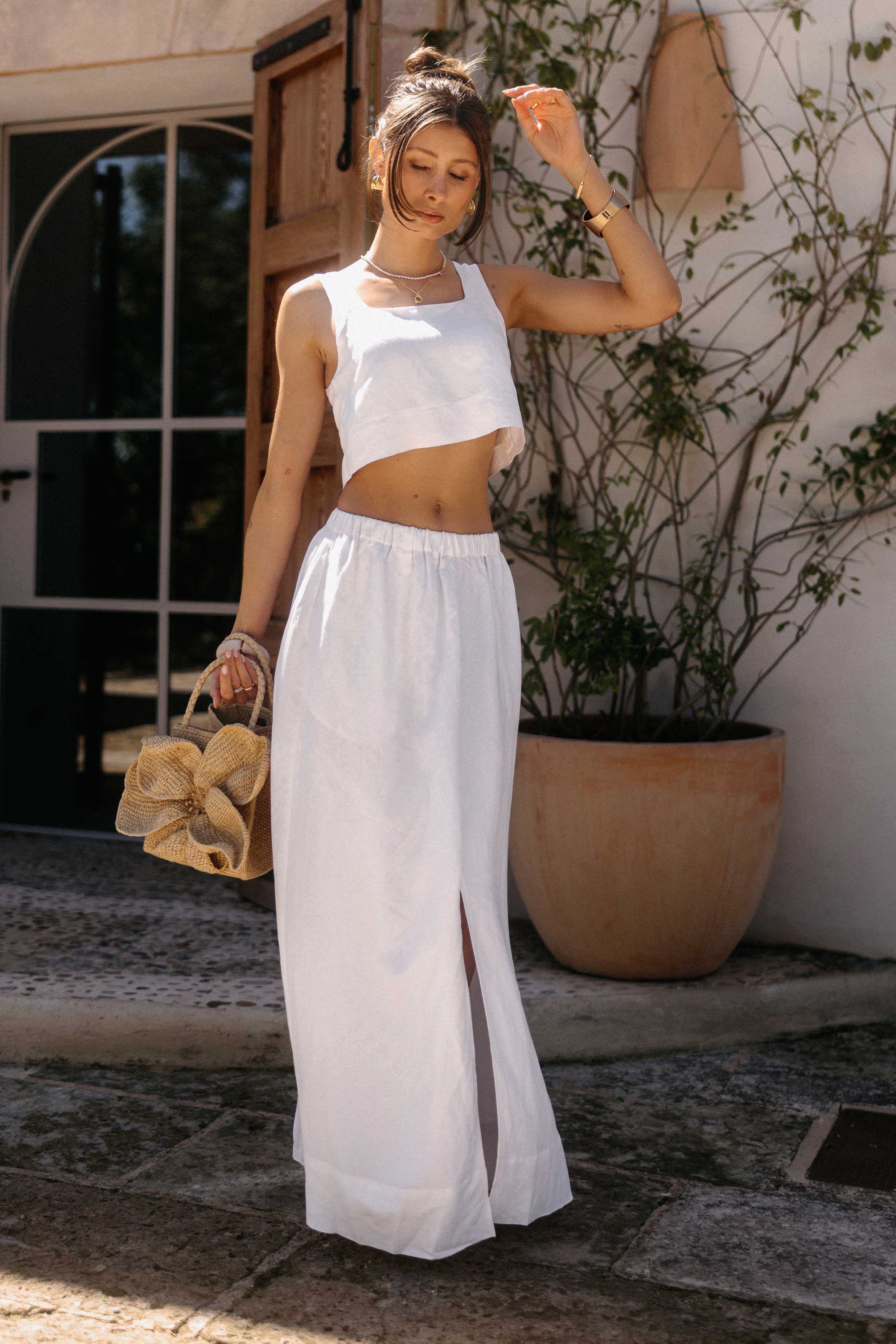 Frau in einem weißen Zweiteiler aus ärmellosem Crop-Top und langem, fließendem Rock läuft durch einen mediterranen Innenhof. In der Hand trägt sie eine geflochtene Strohtasche mit großer Blütenapplikation. Warme Sonnenstrahlen, offene Holztüren und große Pflanzgefäße verleihen der Szene eine sommerlich-elegante Atmosphäre.