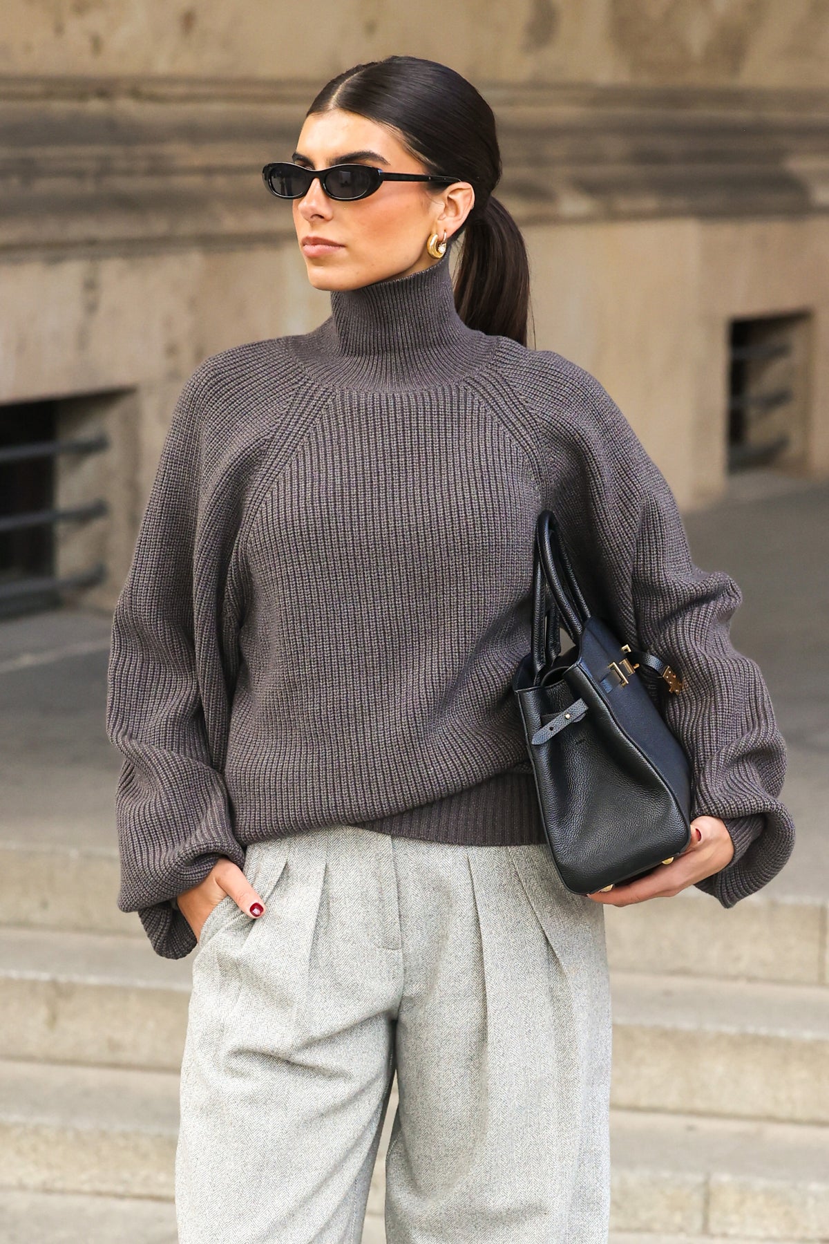 Frau mit dunklem Zopf und schwarzer Sonnenbrille trägt einen oversized Rollkragenpullover in Dunkelgrau, eine graue Bundfaltenhose und eine schwarze Ledertasche vor einer Steinfassade