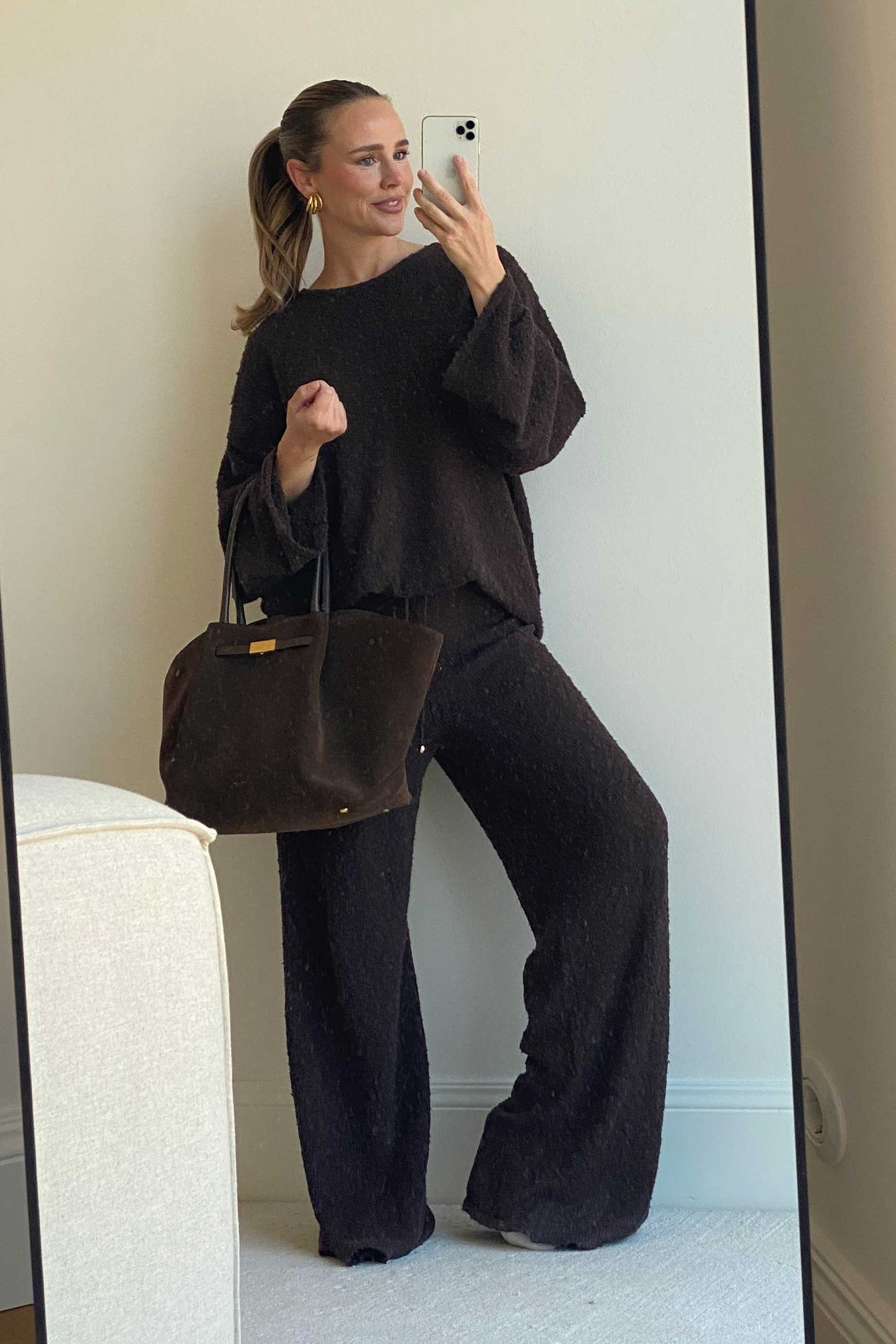 Frau fotografiert sich im Spiegel, trägt ein gemütliches, lockeres Strick-Ensemble in dunklem Braun, bestehend aus weitem Pullover und passender Hose. Dazu kombiniert sie goldene Creolen und eine große, strukturierte Stofftasche in Braun. Sie steht in einem hellen Raum auf einem Teppichboden und wirkt entspannt und stilvoll.