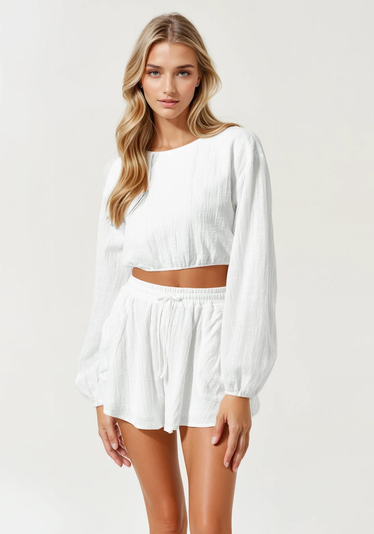 Model im weißen Sommer-Zweiteiler mit bauchfreiem Langarm-Top und Shorts mit Bindeband, aufgenommen aus mittlerer Distanz.