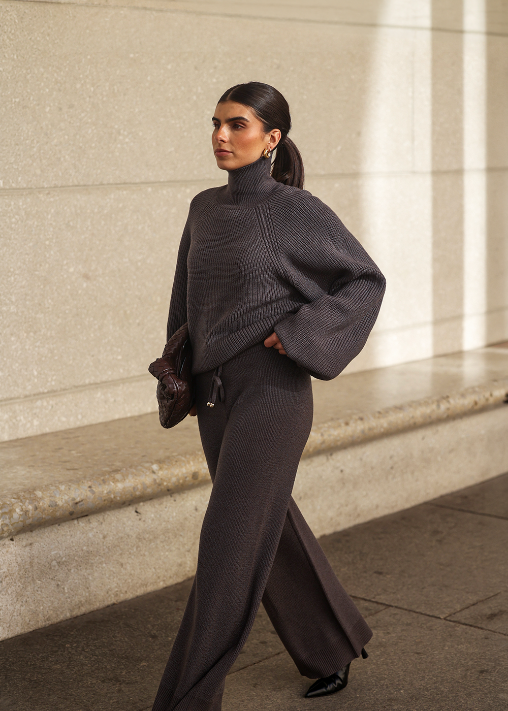 Frau mit dunklem Pferdeschwanz und goldenen Ohrringen läuft in einem dunkelgrauen Strick-Set aus oversized Rollkragenpullover und weiter Strickhose, dazu schwarze Spitzschuhe und eine braune Clutch, vor heller Steinwand im Sonnenlicht