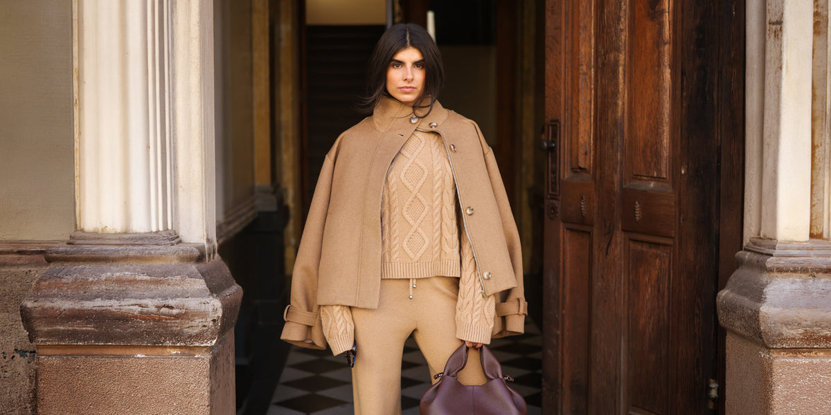 Model in einem monochromen beigefarbenen Outfit aus Cape-Jacke, Zopfmuster-Strickpullover und Strickhose vor einem Hauseingang.