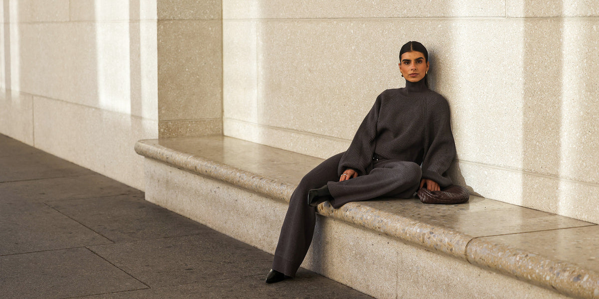 Frau mit zurückgebundenem dunklem Haar lehnt entspannt an einer hellen Steinwand und sitzt auf einer Steinbank, gekleidet in ein dunkelgraues Strick-Set aus oversized Rollkragenpullover und weiter Strickhose, mit schwarzen Spitzschuhen und brauner Clutch, im warmen Sonnenlicht