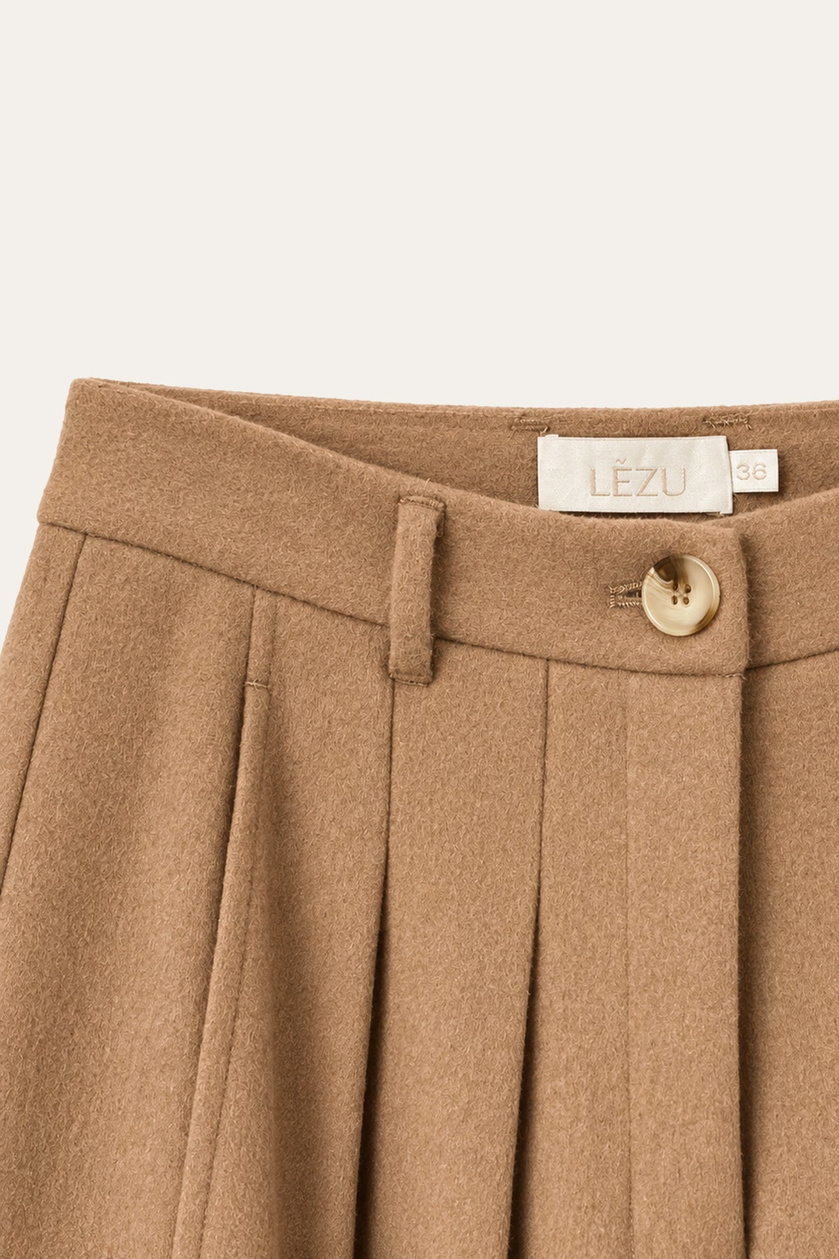 Detailansicht einer camelbraunen Hose mit Bundfalten, Gürtelschlaufen, braun-beigem Knopf und LEZU-Etikett am Bund.