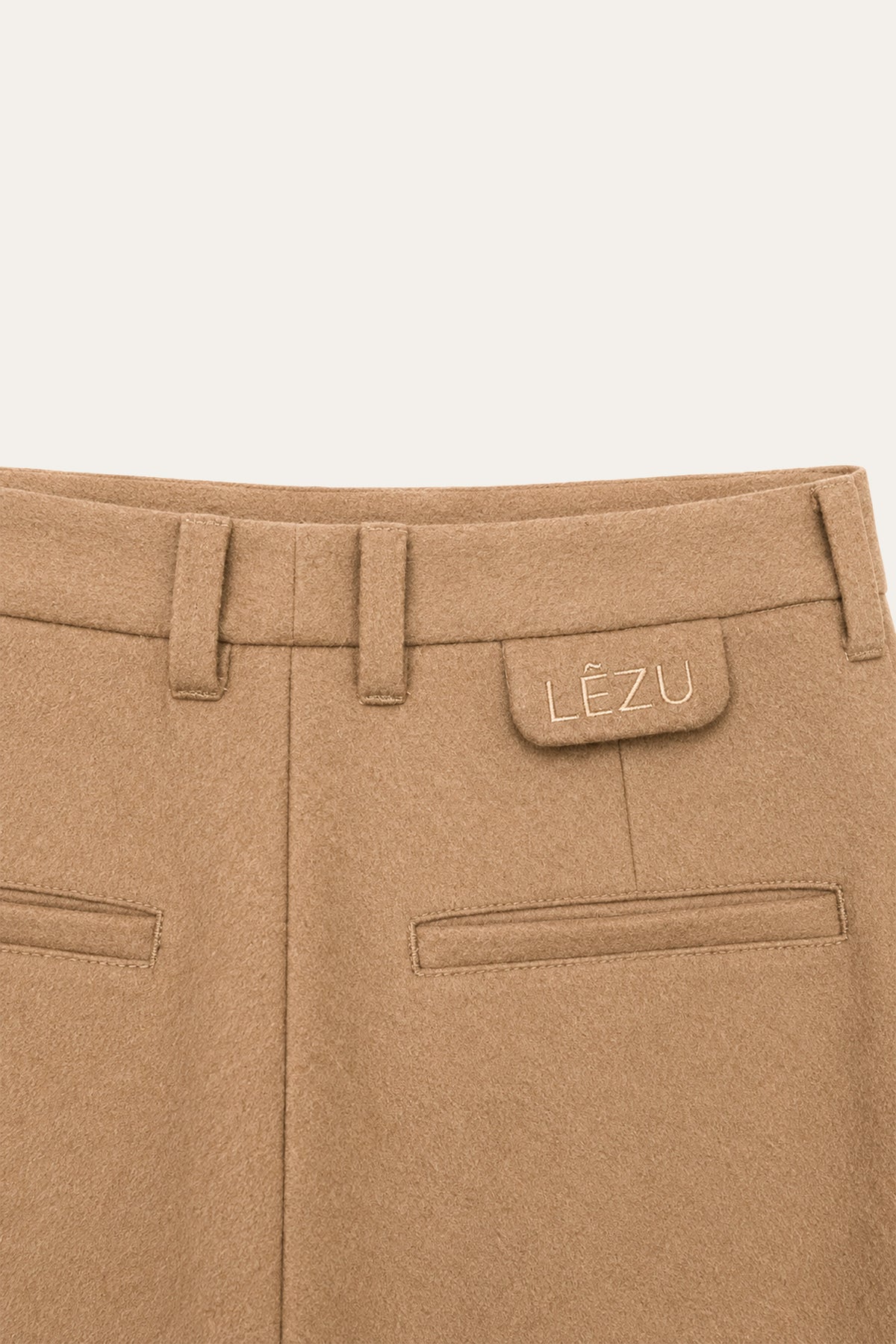 Detailansicht der Rückseite einer camelbraunen Hose mit Gürtelschlaufen, zwei Leistentaschen und LEZU-Stickerei am Bund.