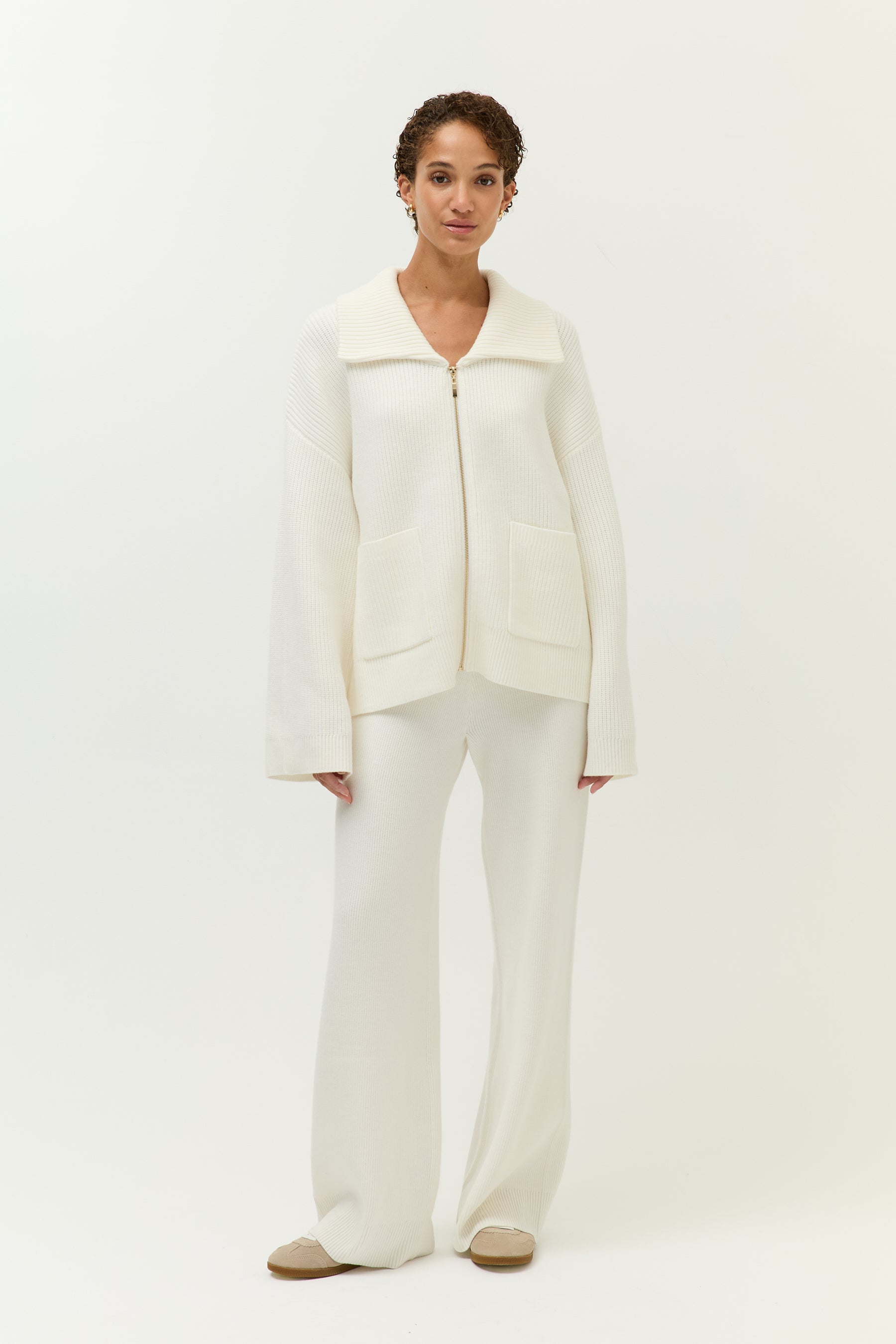 Look 2 – Strickset in Off-White (Bilder 7–12):
Modell trägt ein zweiteiliges Set aus weichem, geripptem Strick in Off-White, bestehend aus einem langärmeligen Zip-Cardigan mit breitem Kragen, goldfarbenem Reißverschluss und zwei großen aufgesetzten Taschen sowie einer weit geschnittenen Hose mit fließendem Fall. Auf der Rückseite der Jacke ist ein kleines, goldfarbenes Lezu-Logo unter dem Kragen platziert. Styling mit beigen Slippern.