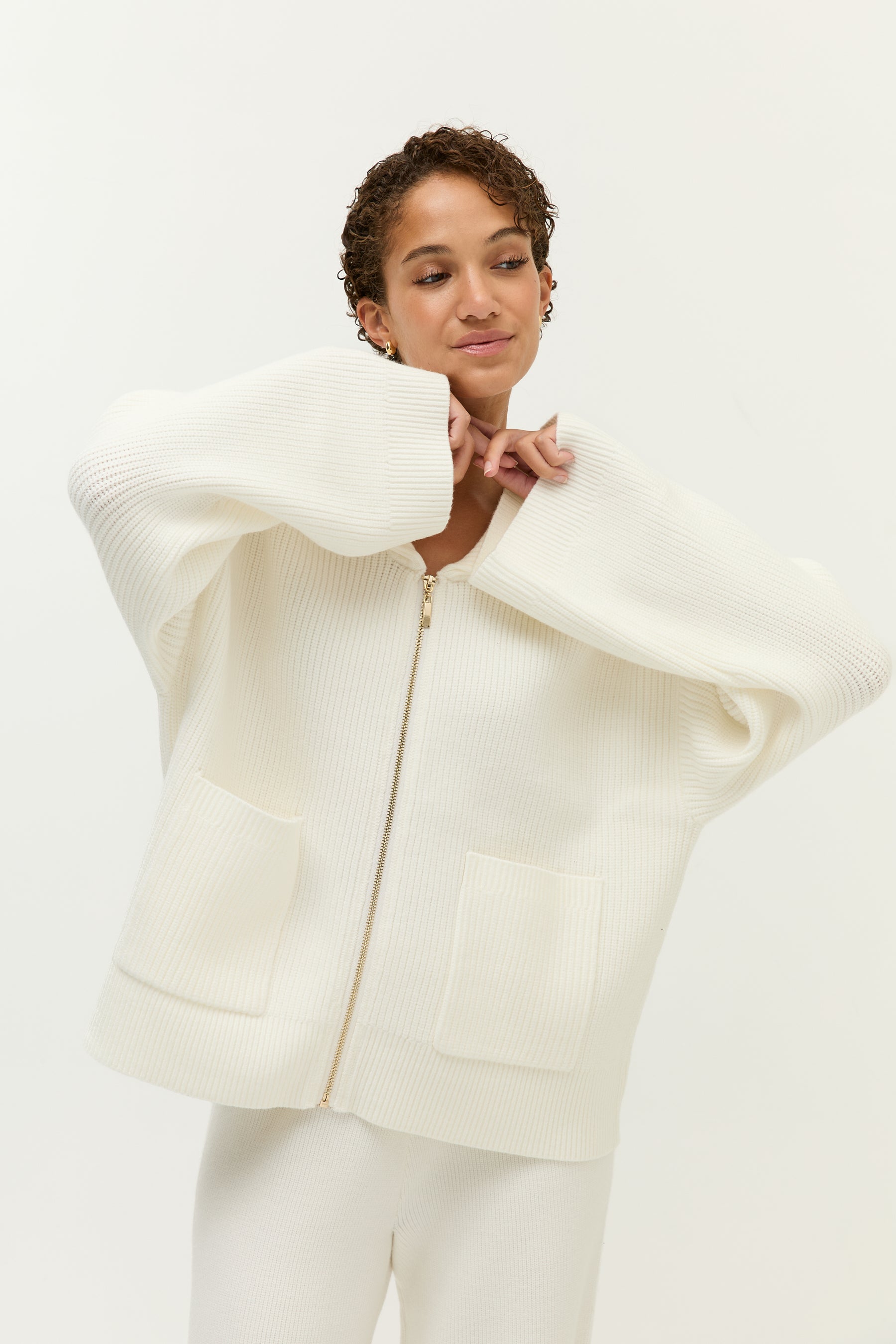 Look 1 – Strickkleid in Beige (Bilder 1–6):
Modell trägt ein beiges, grob gestricktes Minikleid mit langen Ärmeln, gerippten Bündchen und zwei aufgesetzten Brusttaschen mit goldfarbenen Zierknöpfen. Das Kleid hat einen gerippten Rundhalsausschnitt und fällt locker über den Körper. Die Rückseite zeigt ein kleines, goldfarbenes Lezu-Logo unter dem Kragen. Styling mit dunklen Mokassins und beigen Socken.