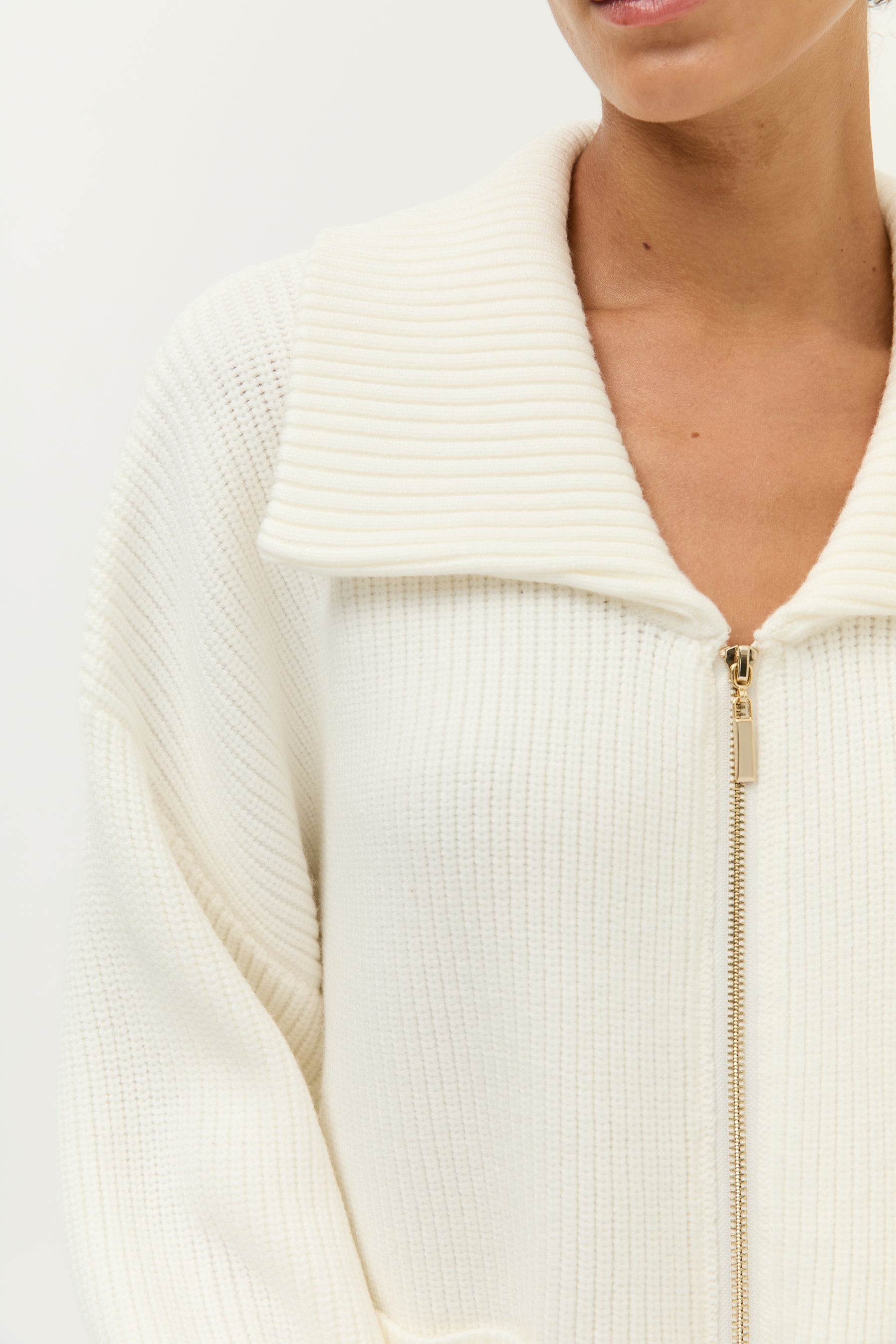 Look 2 – Strickset in Off-White (Bilder 7–12):
Modell trägt ein zweiteiliges Set aus weichem, geripptem Strick in Off-White, bestehend aus einem langärmeligen Zip-Cardigan mit breitem Kragen, goldfarbenem Reißverschluss und zwei großen aufgesetzten Taschen sowie einer weit geschnittenen Hose mit fließendem Fall. Auf der Rückseite der Jacke ist ein kleines, goldfarbenes Lezu-Logo unter dem Kragen platziert. Styling mit beigen Slippern.