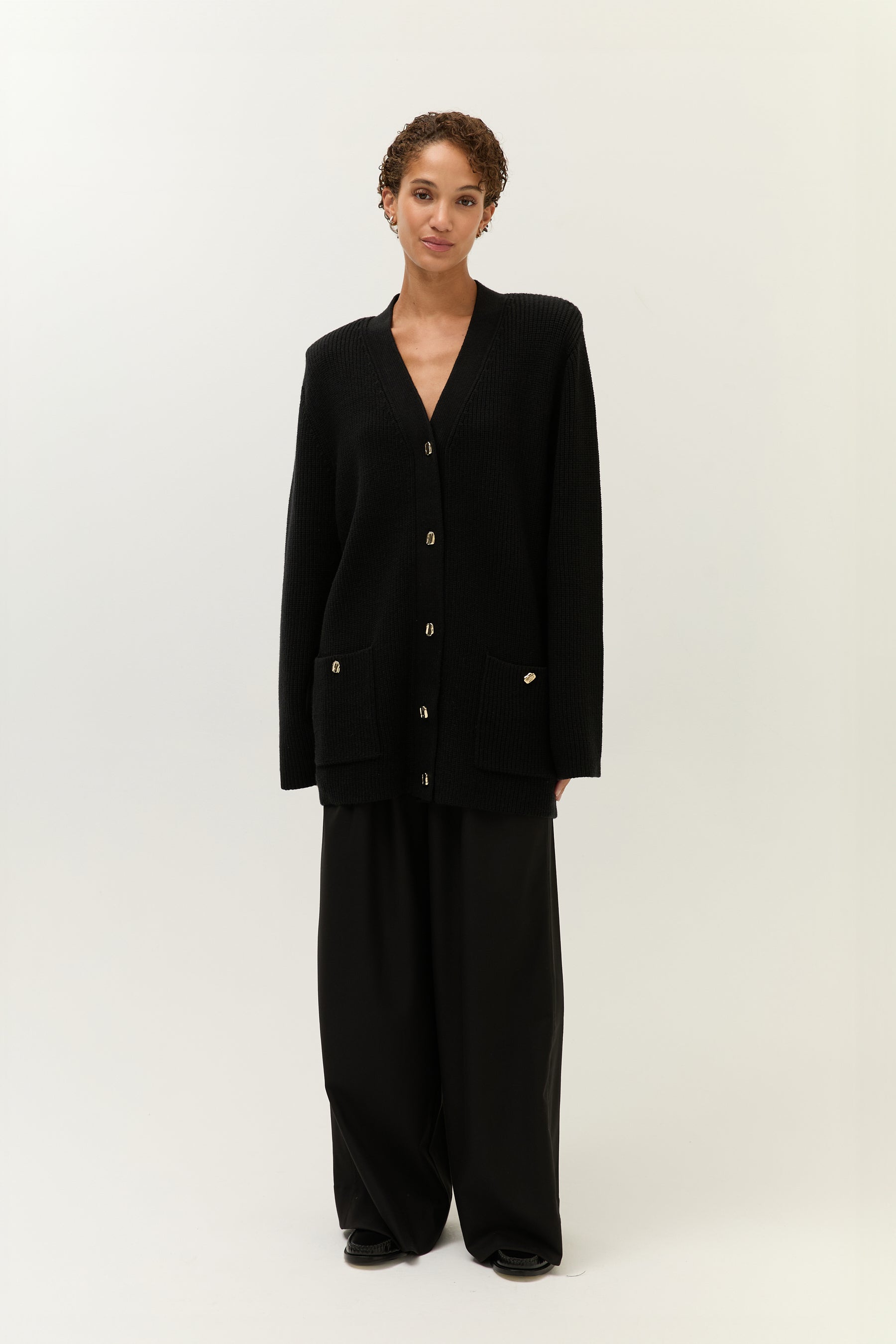 Model in einem langen, schwarzen Strick-Cardigan mit V-Ausschnitt, goldenen Knöpfen und aufgesetzten Taschen. Dazu trägt sie eine weite, schwarze Hose und schwarze Loafer.
