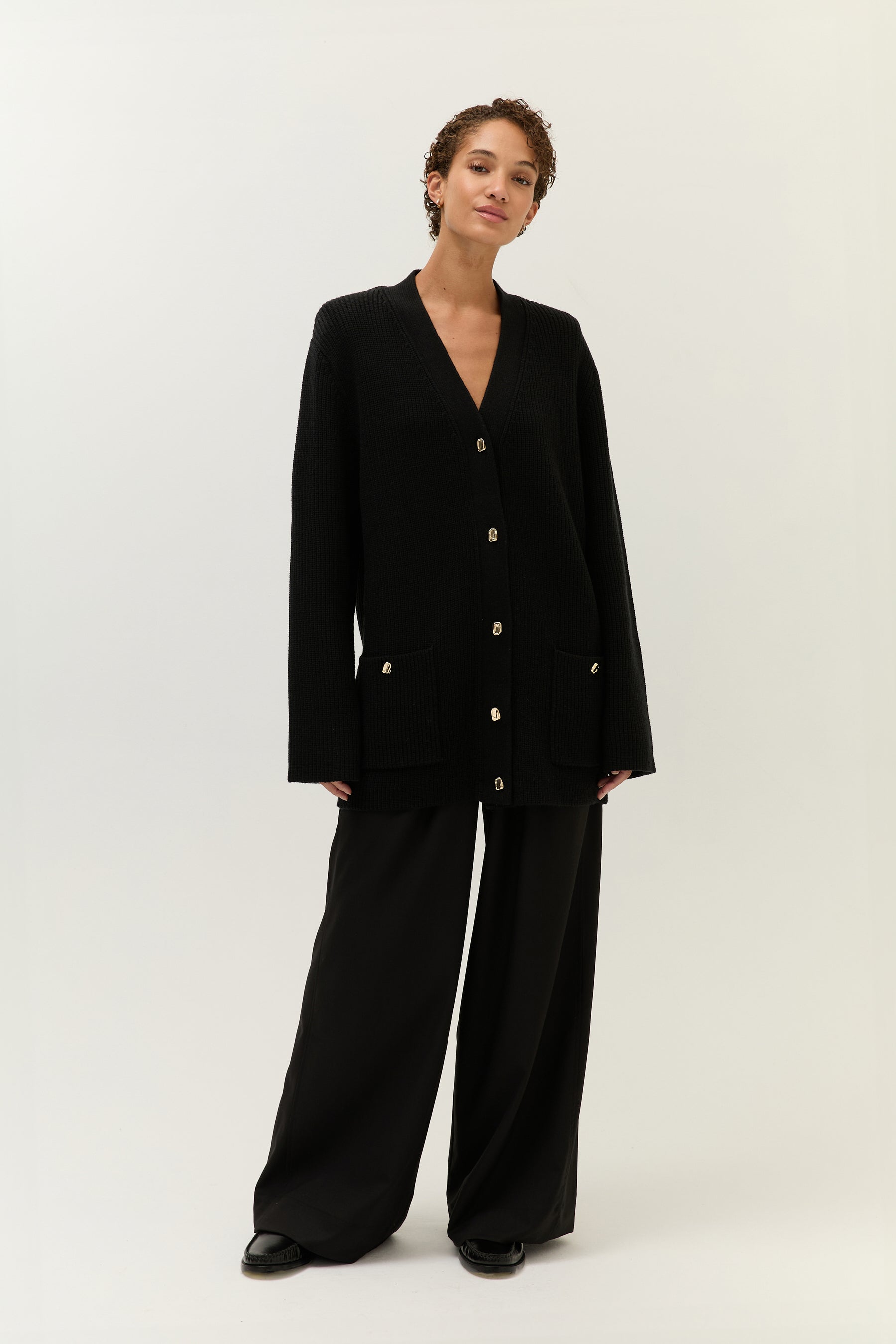 Frontalansicht des Models in einem schwarzen Strick-Cardigan mit goldenen Knöpfen, kombiniert mit einer weiten schwarzen Hose.