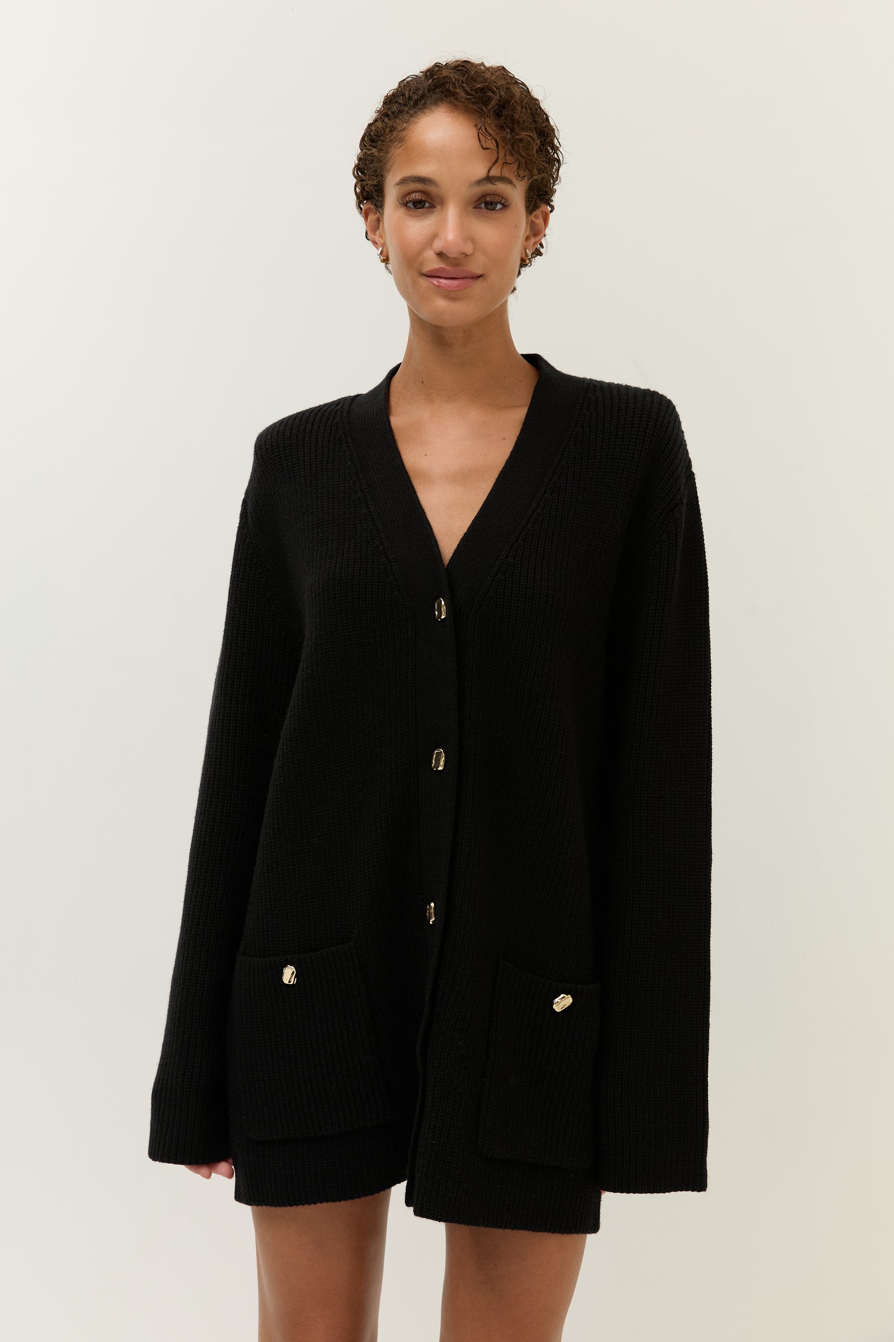 Halbnahe Frontaufnahme des Models, das den schwarzen Strick-Cardigan ohne Hose oder mit Shorts darunter trägt. Der Fokus liegt auf dem Oberteil und dem Ausschnitt.