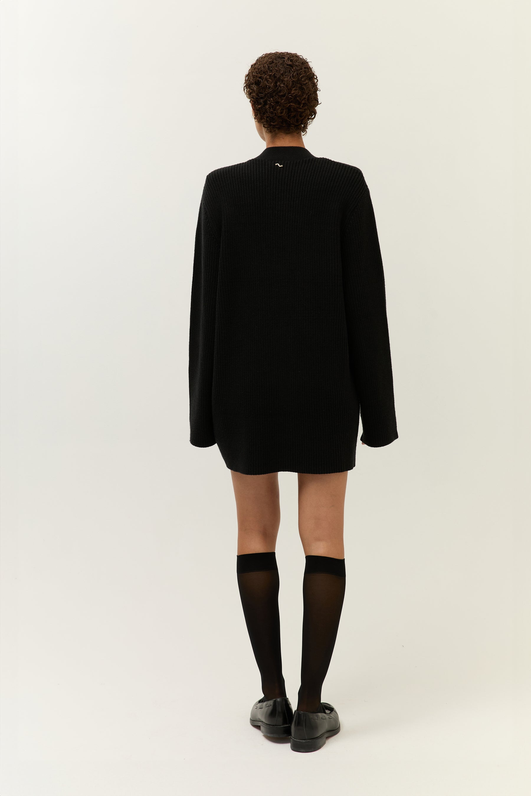Rückansicht des Models, das den Cardigan wie ein Minikleid trägt, kombiniert mit knielangen, transparenten Strümpfen und schwarzen Loafern.