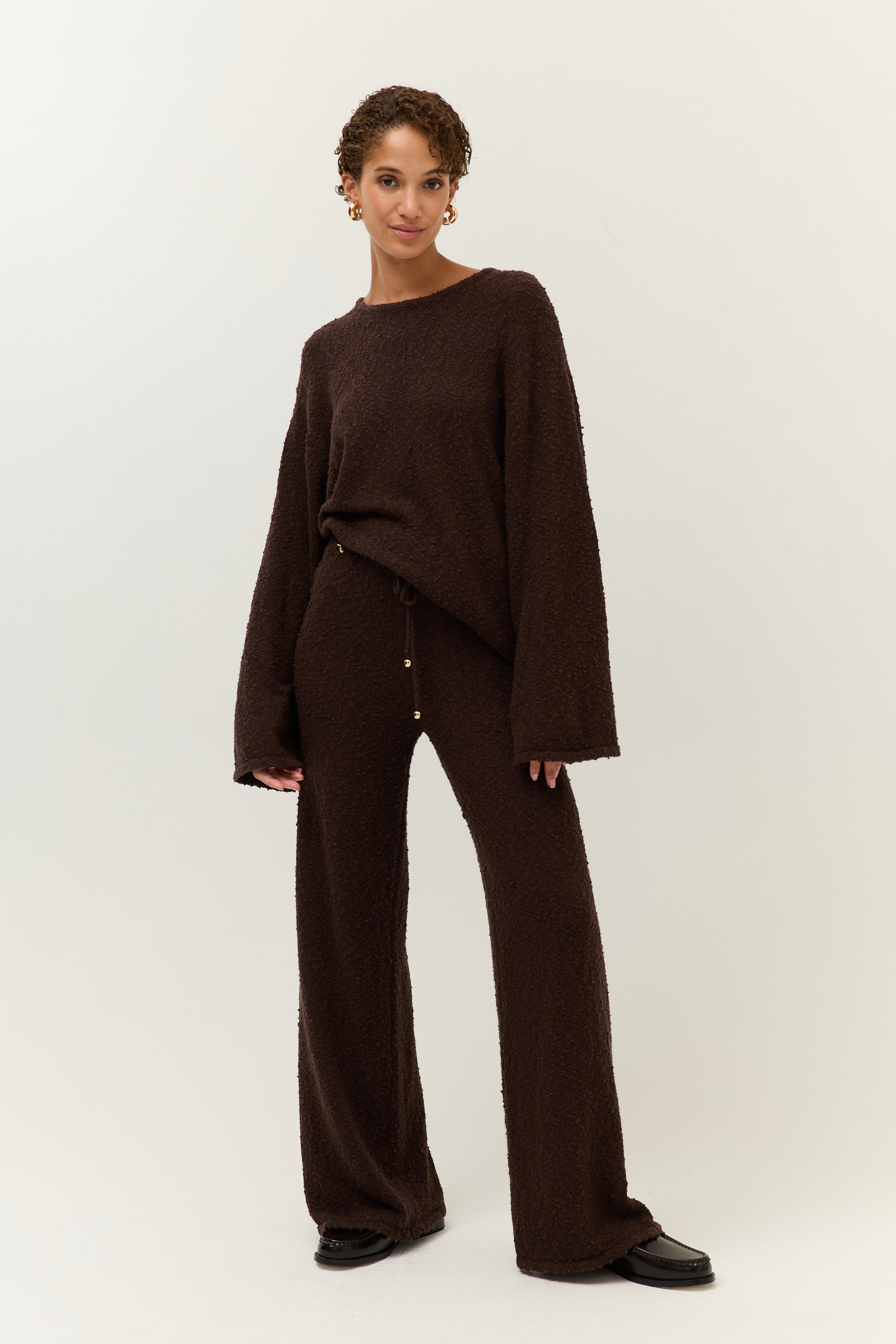 Ganzkörperaufnahme: Model trägt ein weites braunes Bouclé-Set aus Pullover und Hose mit Kordelzug vor hellem Hintergrund.