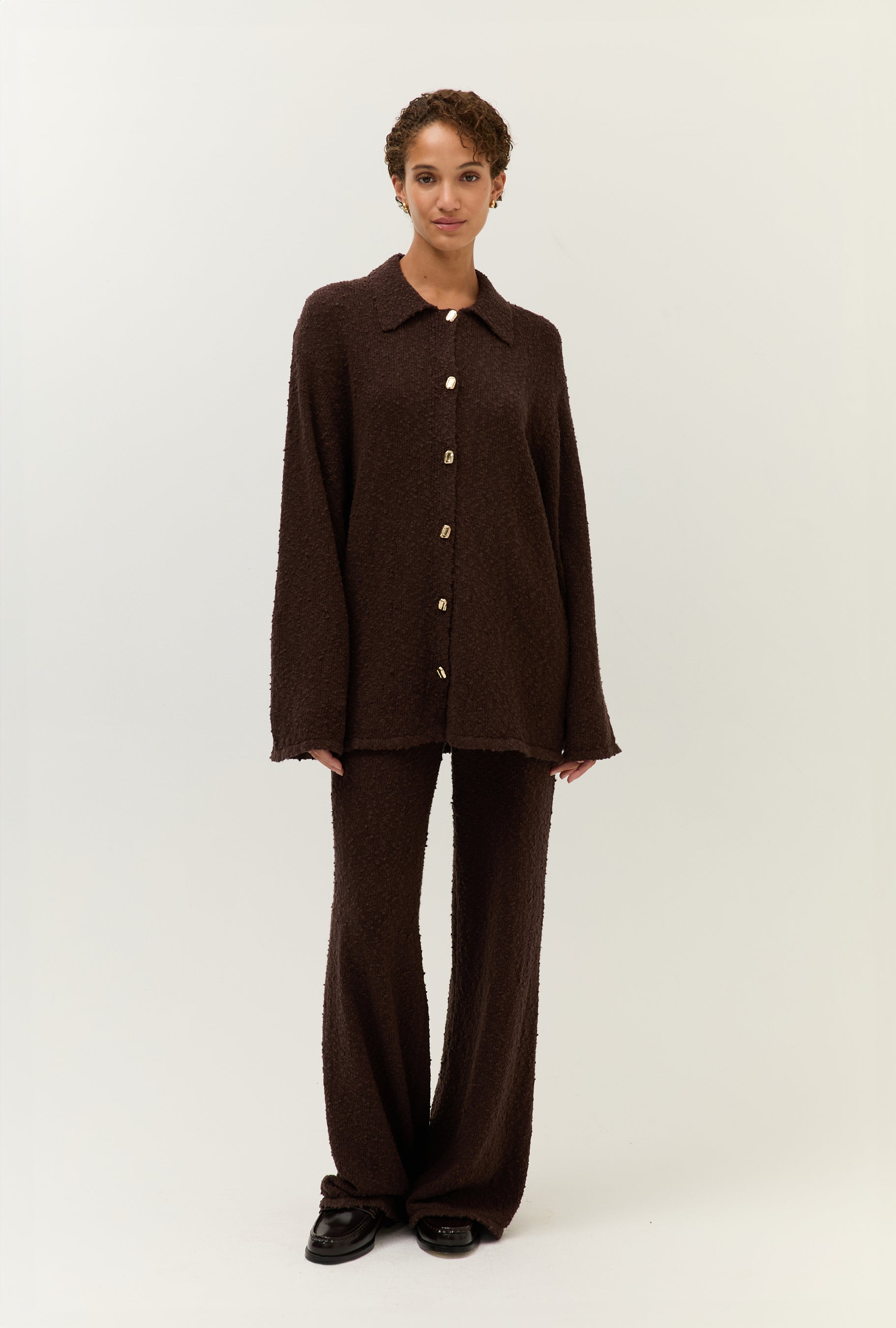 Das Model trägt ein braunes zweiteiliges Set aus strukturierter Bouclé-Ware, bestehend aus einem locker geschnittenen Hemd mit goldenen Knöpfen und einer weiten Hose.