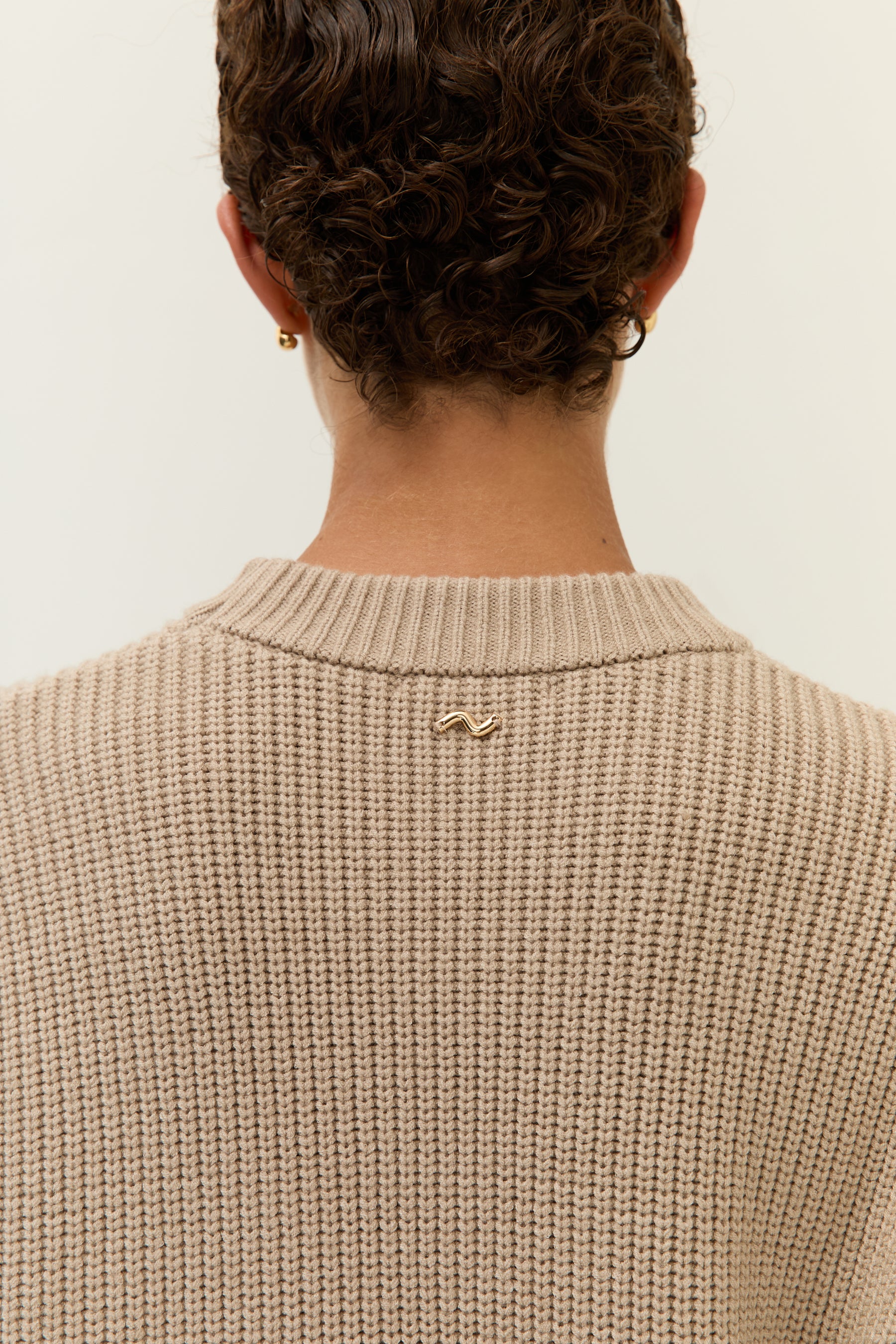 Rückansicht des Kragens mit Schmuckdetail
Detailaufnahme vom Nackenbereich des Strickkleids mit kleinem goldenen Logo-Emblem unter dem Kragen.