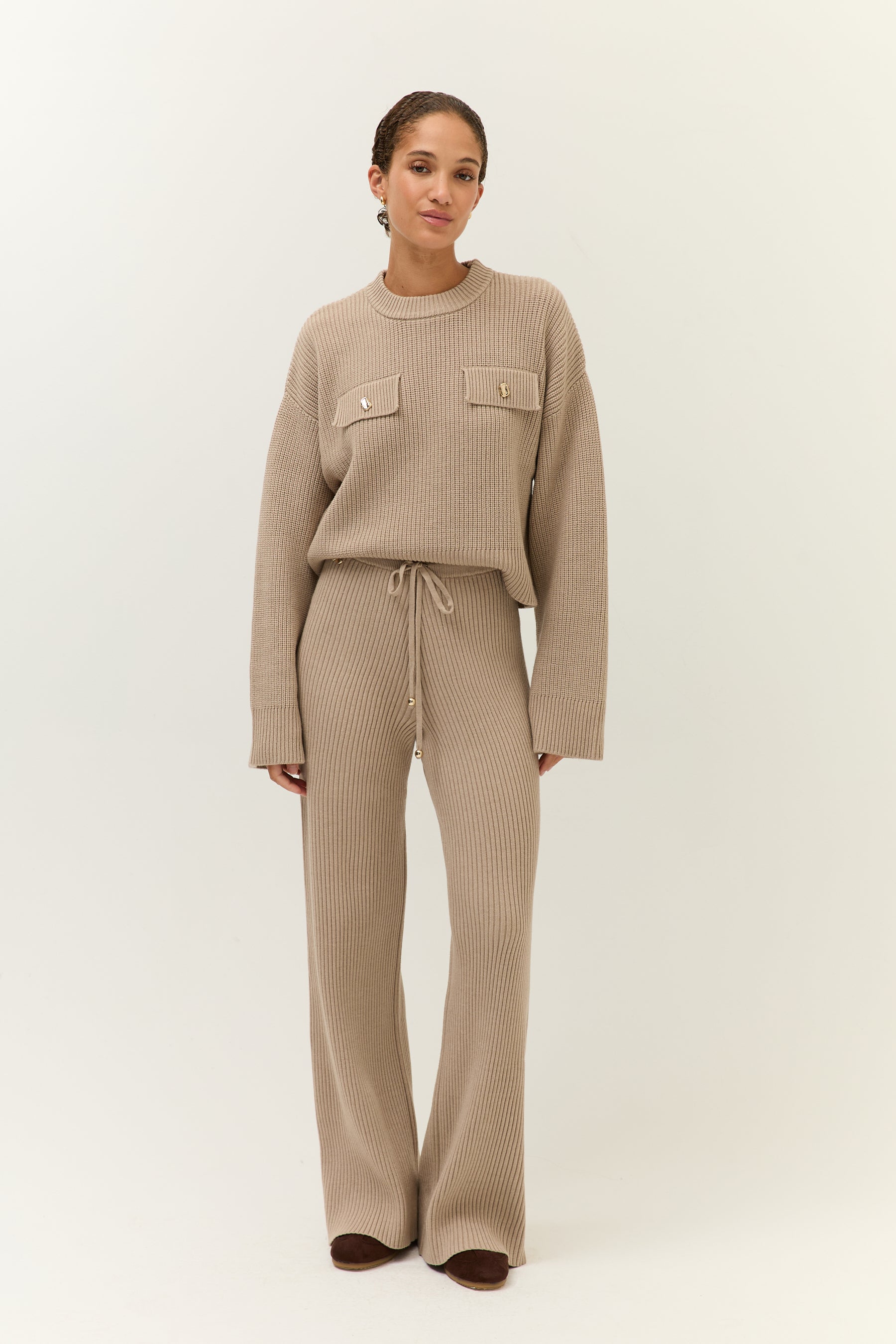 Model trägt ein beiges Strick-Ensemble mit weitem Pullover und passender Hose in gerippter Optik, frontal fotografiert.
