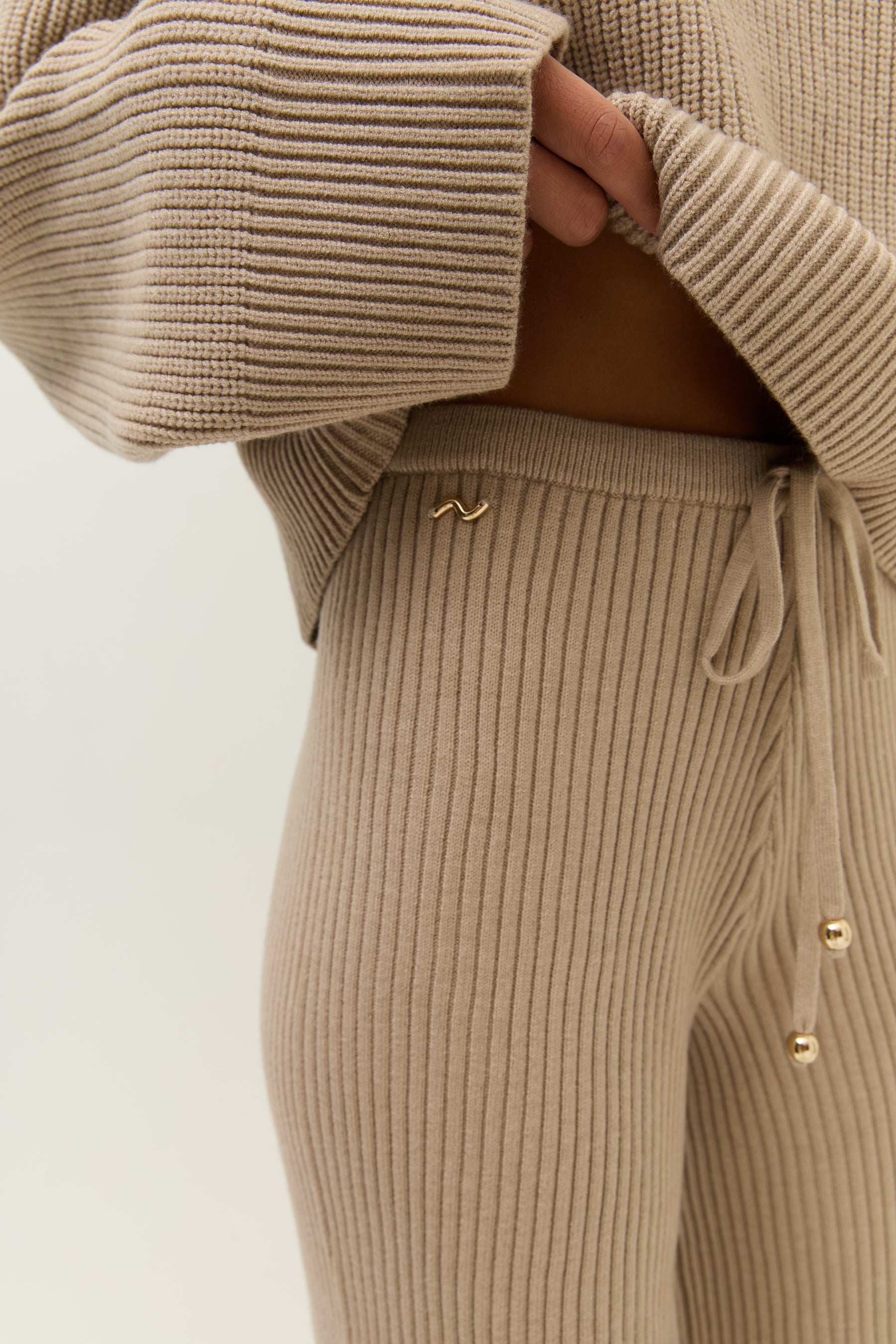 Makroaufnahme des goldenen Logo-Pins auf dem Hosenbund, eingerahmt vom gerippten Strickstoff.