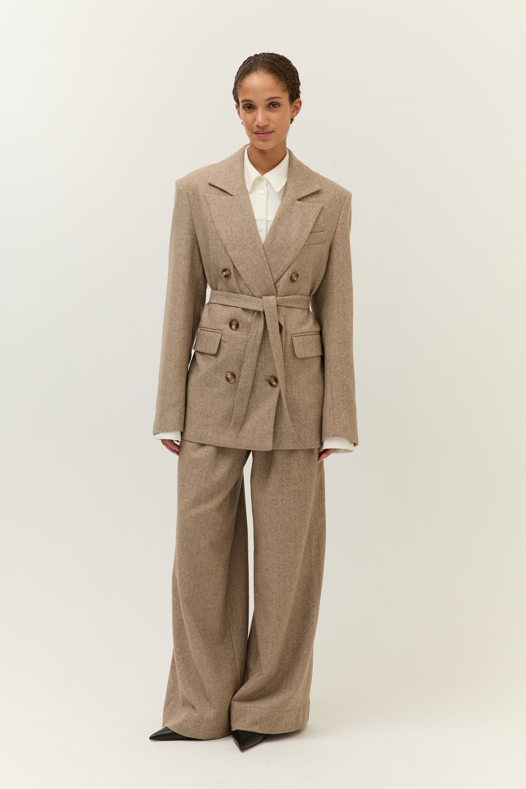 Look mit geschlossenem Gürtel-Blazer
Model trägt einen doppelreihigen Blazer mit Bindegürtel und passender weiter Hose in Hellbraun. Darunter ein weißes Hemd mit Kragen.