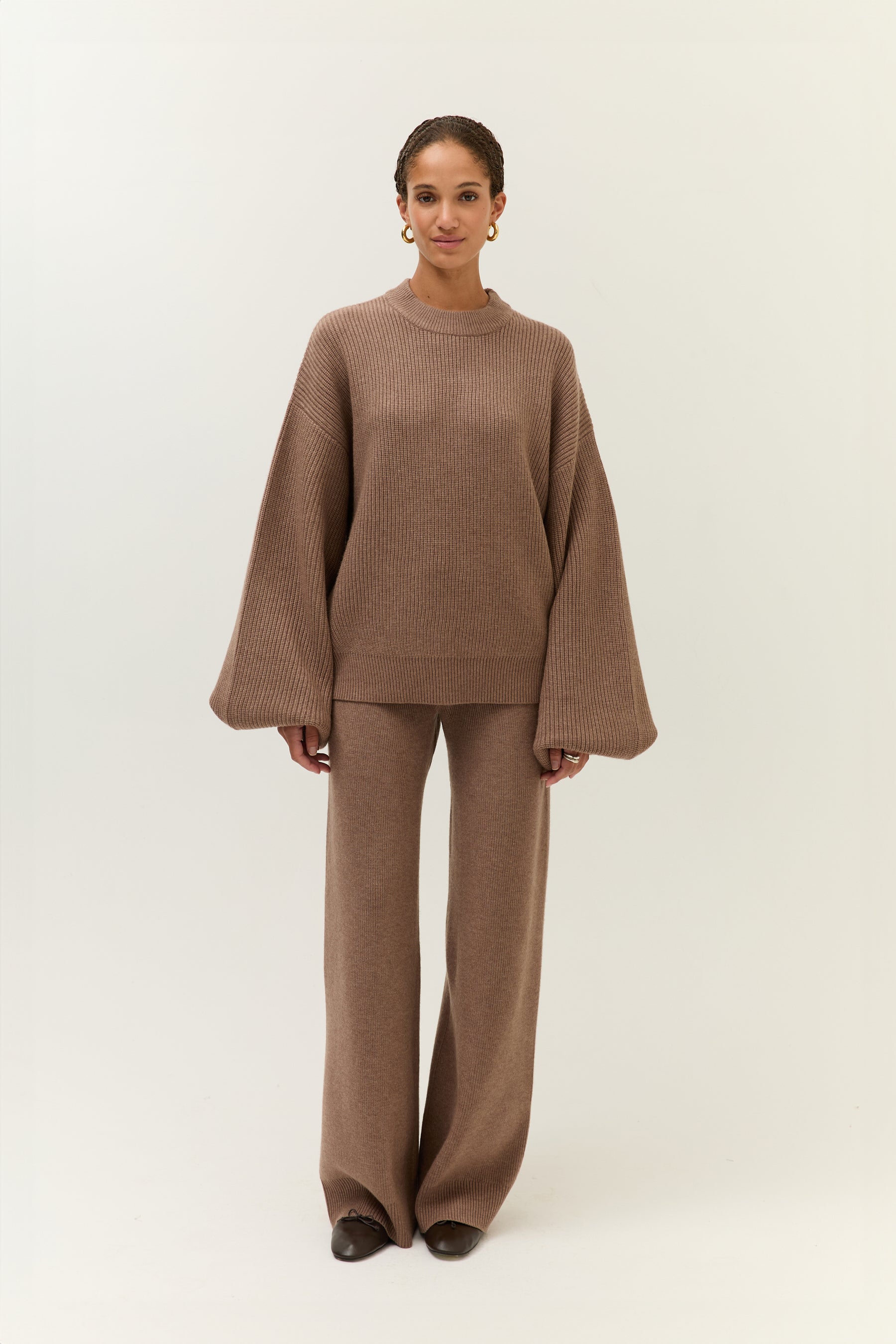 rontalansicht des vollständigen Looks. Das Modell steht gerade, trägt das Set aus locker fallendem Pullover mit weiten Ärmeln und einer weit geschnittenen Hose aus weichem, braunem Strick.