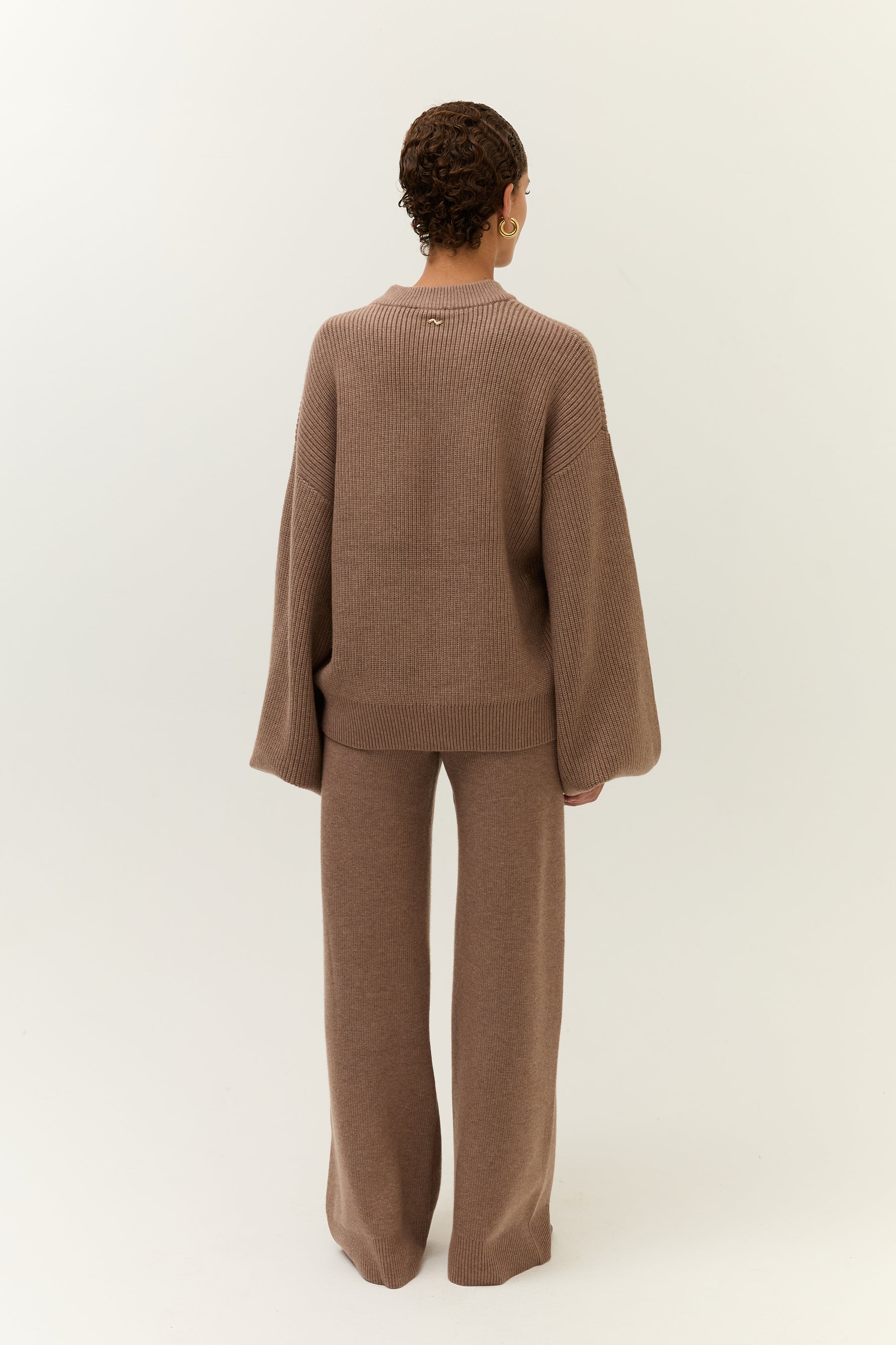 Rückansicht des Modells, das den Pullover mit einer Hand locker anhebt. Der Schnitt der Hose ist betont figurumschmeichelnd.