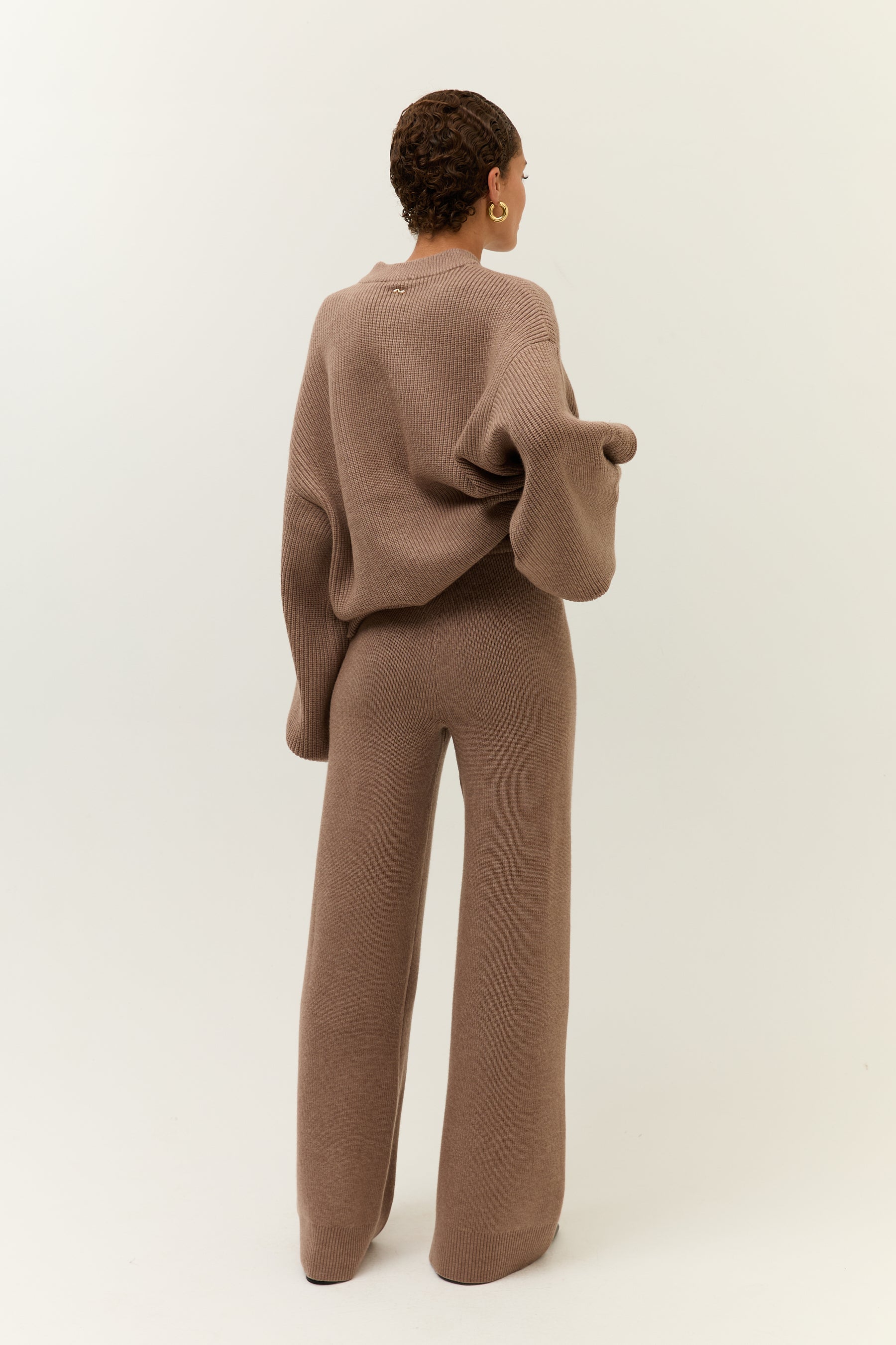 Rückansicht des Modells, das den Pullover mit einer Hand locker anhebt. Der Schnitt der Hose ist betont figurumschmeichelnd.