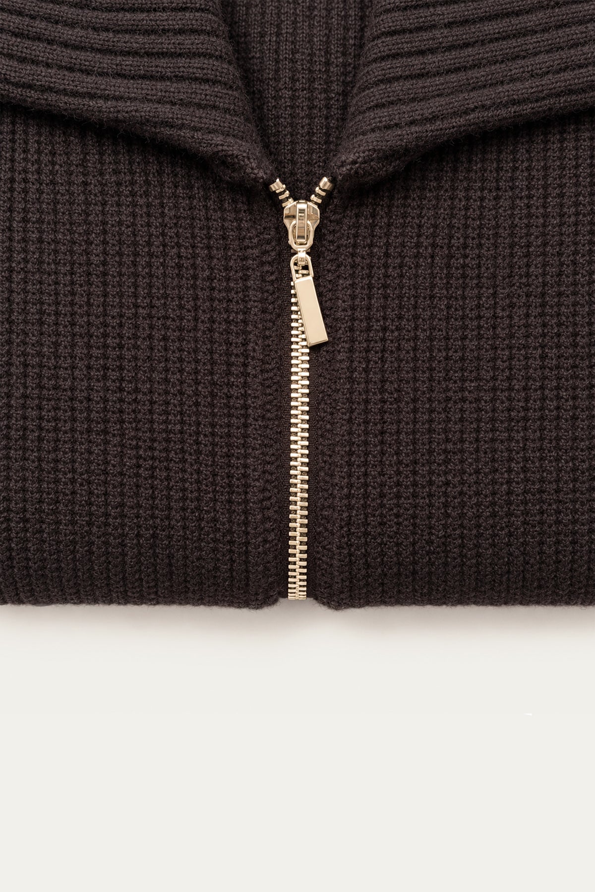 Detailaufnahme eines dunkelbraunen Strickoberteils mit geripptem Kragen und goldenem Reißverschluss auf weißem Hintergrund.
