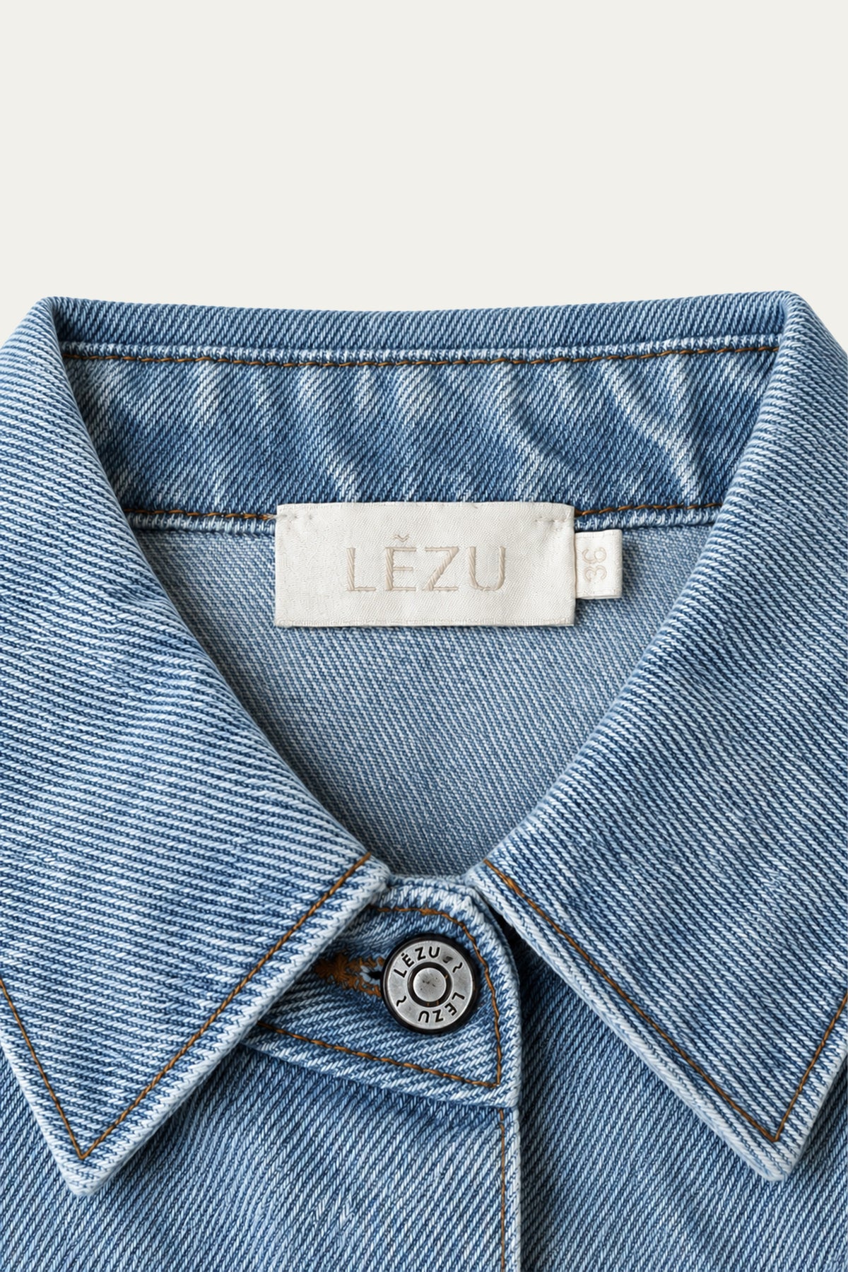 Detailansicht eines hellblauen Jeanshemds mit spitzem Kragen, kontrastierenden Nähten, Metallknopf mit LEZU-Schriftzug und LEZU-Label am Innenkragen.c