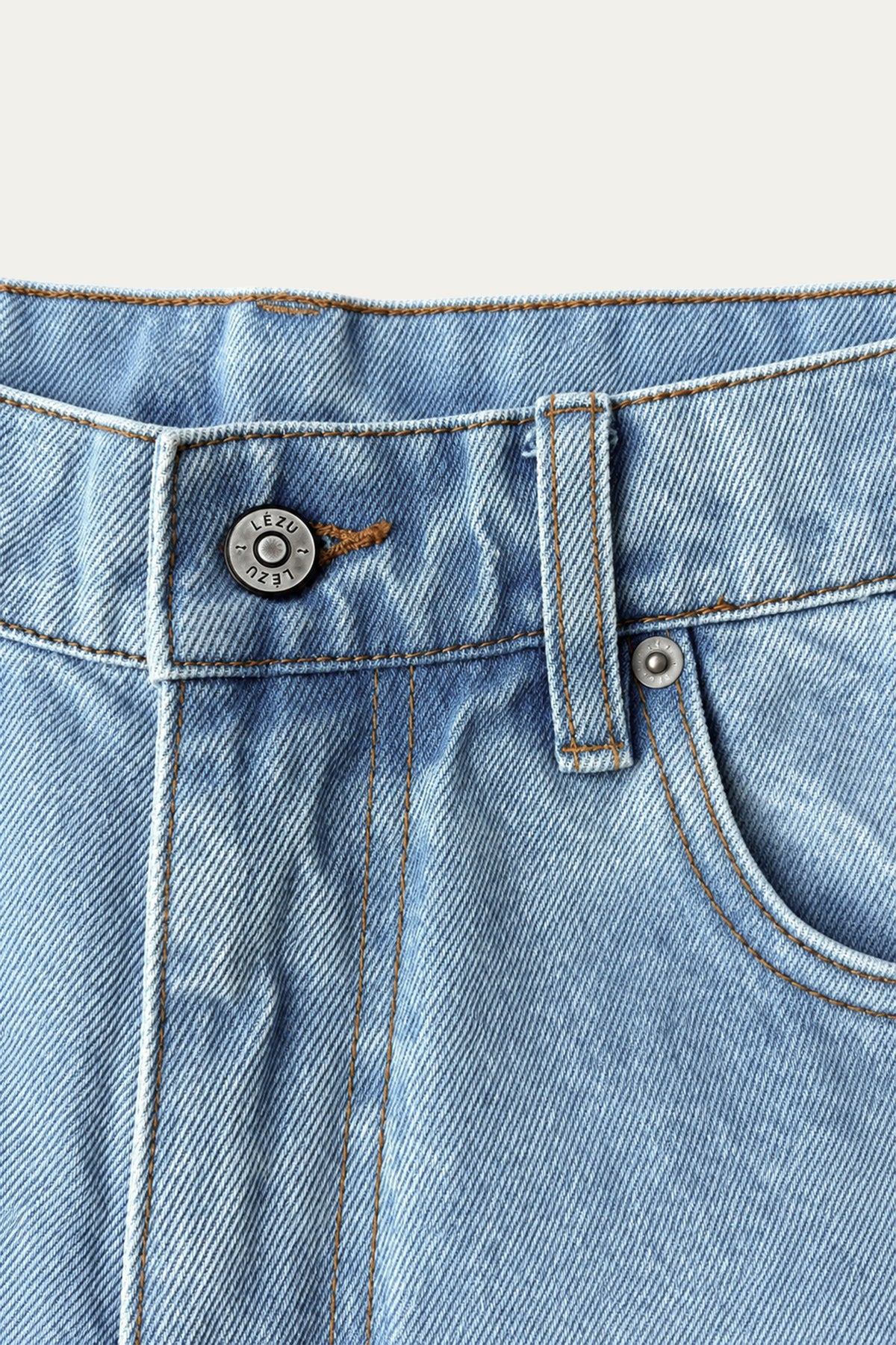 Detailansicht der Vorderseite einer hellblauen Jeans mit Bund, Gürtelschlaufe, Eingrifftasche und Metallknopf mit LEZU-Schriftzug.