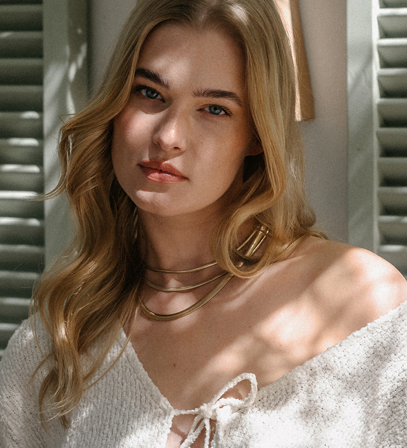 Close-up-Porträt einer blonden Frau mit gewelltem Haar, die eine markante, mehrlagige, starre goldene Gliederkette trägt. Sie blickt direkt in die Kamera und trägt ein helles, off-shoulder Oberteil mit Bindedetail. Das Licht fällt durch Zweige und erzeugt ein Spiel aus Schatten auf ihrem Gesicht und Dekolleté.