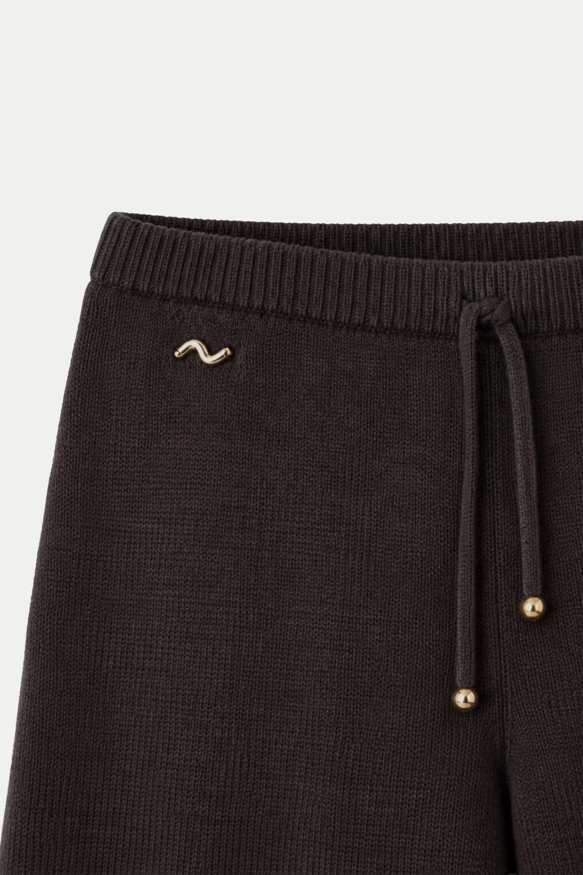 Detailaufnahme einer dunkelbraunen Strickhose mit geripptem Bund, Kordelzug mit goldfarbenen Endkappen und kleinem goldfarbenem Logo.