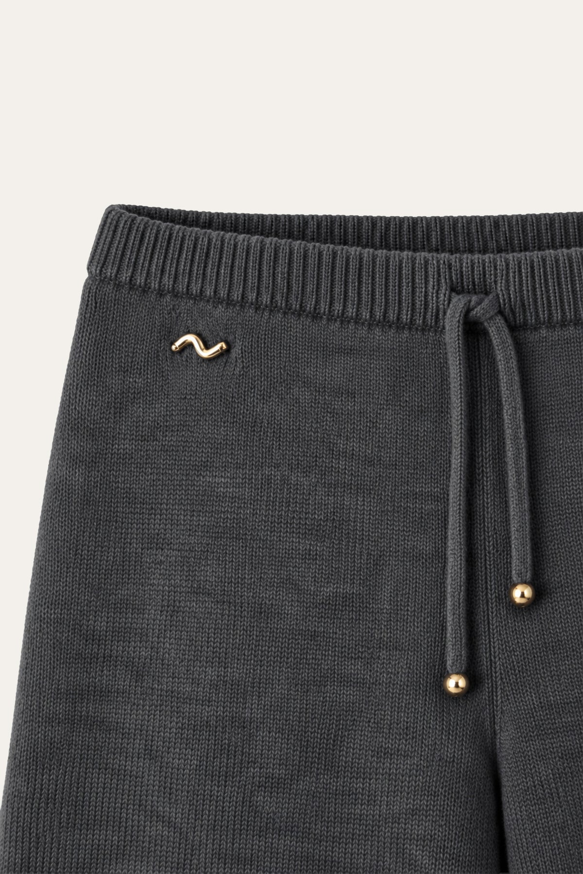 Detailansicht einer dunkelgrauen Strickhose mit geripptem Bund, Kordelzug mit goldfarbenen Enden und goldfarbener Zierapplikation.
