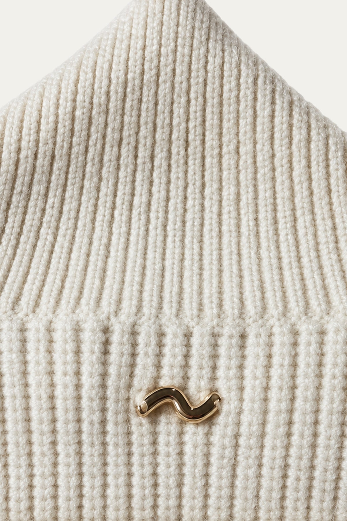 Detailaufnahme eines cremefarbenen Strickpullovers mit Rippenstruktur und kleinem goldfarbenem Logo.