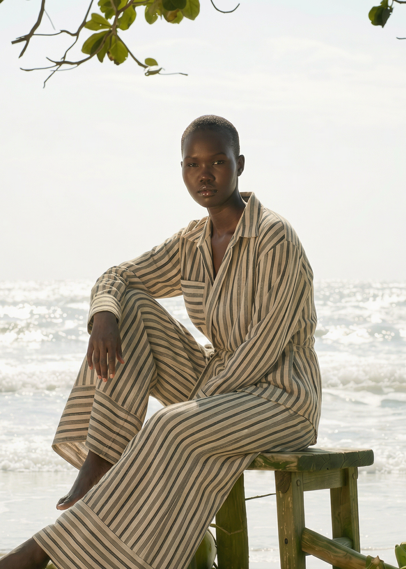 Beige-gestreiftes Zweiteiler-Set aus leichtem Stoff mit locker geschnittenem Hemd und weiter Hose, getragen von einem Model sitzend auf einem Holzstuhl am Strand mit Meerblick im Hintergrund.