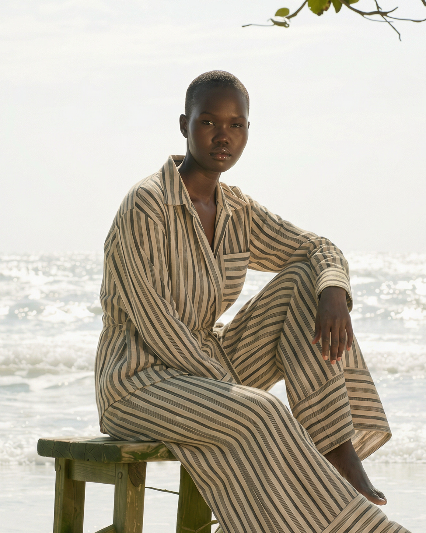 Beige-gestreiftes Zweiteiler-Set aus leichtem Stoff mit locker geschnittenem Hemd und weiter Hose, getragen von einem Model sitzend auf einem Holzstuhl am Strand mit Meerblick im Hintergrund.