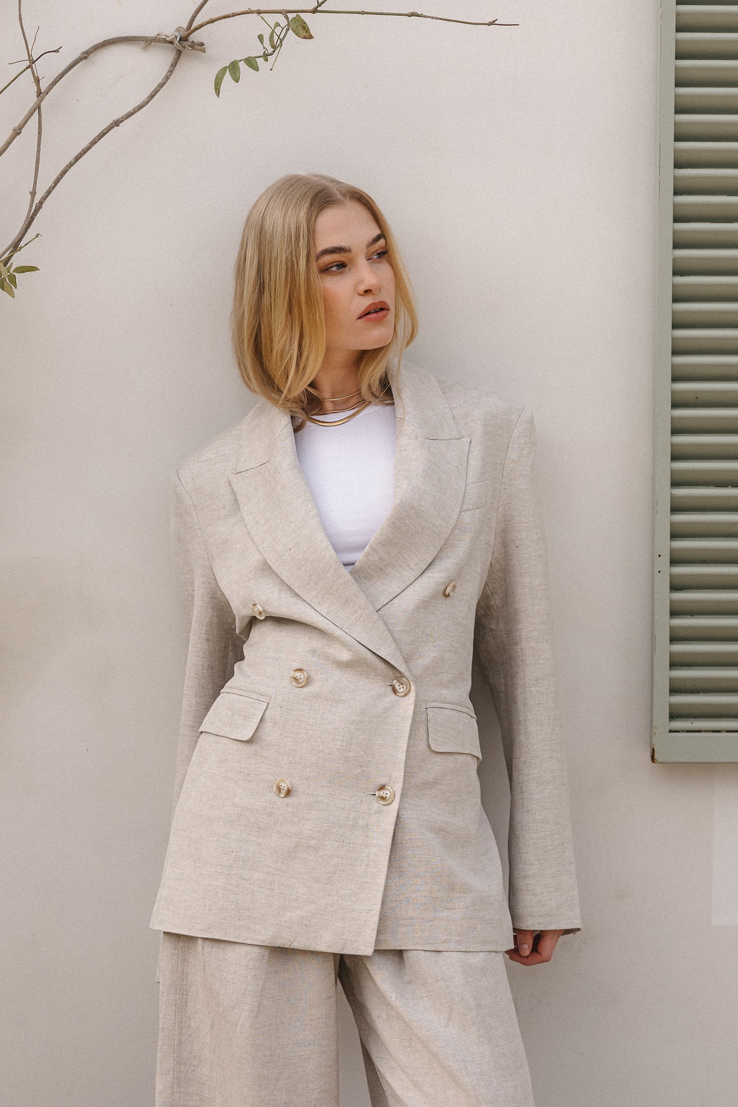 Model trägt ein zweiteiliges beigefarbenes Leinen-Set aus tailliertem doppelreihigem Blazer mit breitem Revers und weiter Hose, kombiniert mit weißem Top, goldfarbenem Halsschmuck und Sandalen