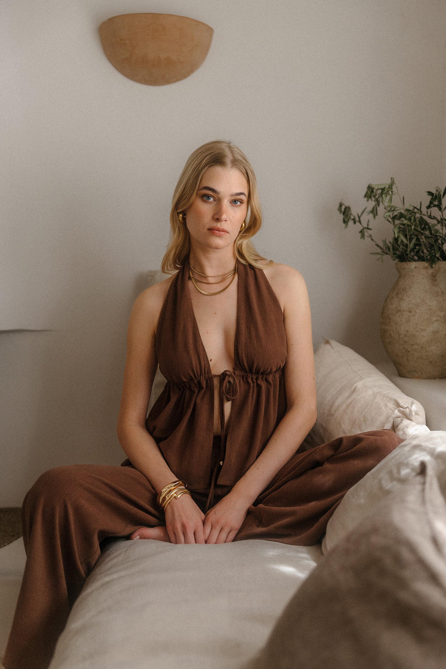 Model sitzt auf einem Sofa und trägt ein braunes, fließendes Haltertop mit Bindeband und tiefem Ausschnitt sowie eine weit geschnittene Hose, kombiniert mit goldenen Armreifen und Kette, vor einer schlichten Wand mit Kissen und Vase