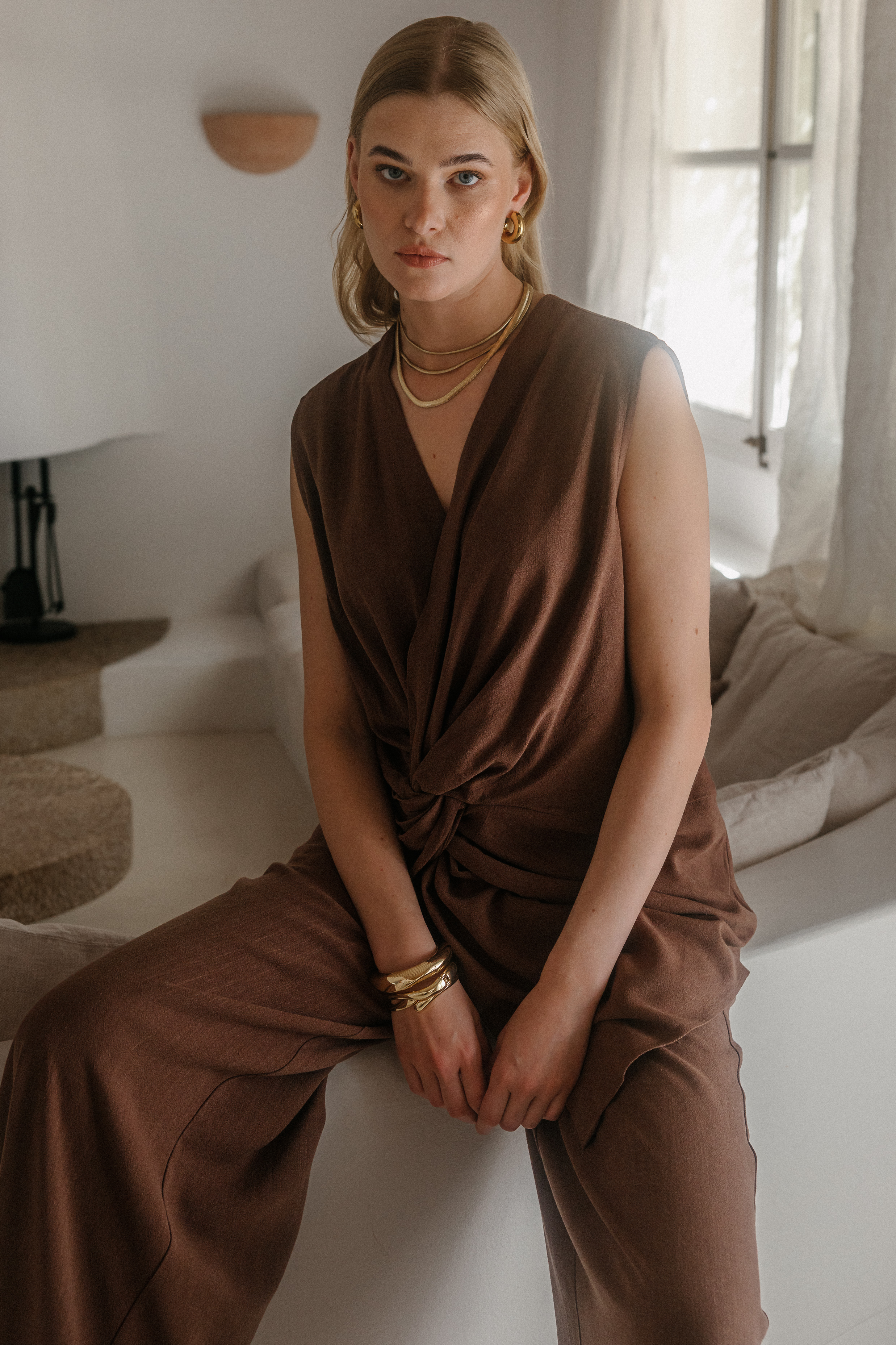 Frau sitzt in einem hellen, minimalistischen Wohnraum und trägt einen ärmellosen braunen Jumpsuit mit drapiertem V-Ausschnitt und weitem Bein. Goldfarbene Schmuckdetails aus Halsreifen, Creolen und Armreifen ergänzen den eleganten Look. Weiches Tageslicht fällt durch die Fenster und unterstreicht die fließende Silhouette des Outfits.