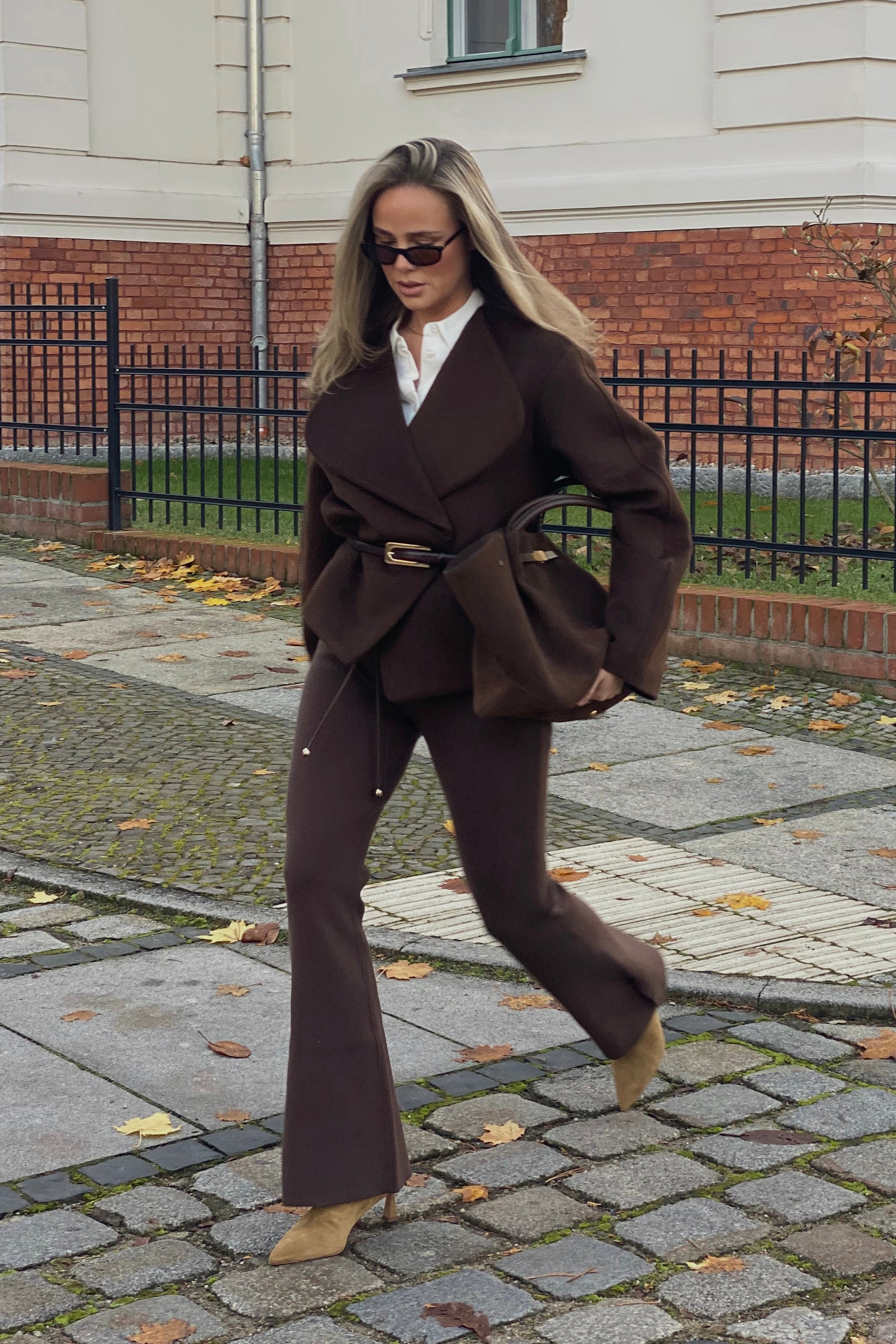 Frau läuft über einen herbstlich bedeckten Gehweg in einem monochromen, braunen Outfit bestehend aus weit geschnittener Jacke mit Taillengürtel und passender Hose mit Schlag. Dazu trägt sie beigefarbene Stiefeletten mit Absatz, eine braune Tasche und eine dunkle Sonnenbrille. Im Hintergrund ist eine Altbaufassade mit Ziegelmauer und schwarzem Zaun zu sehen.