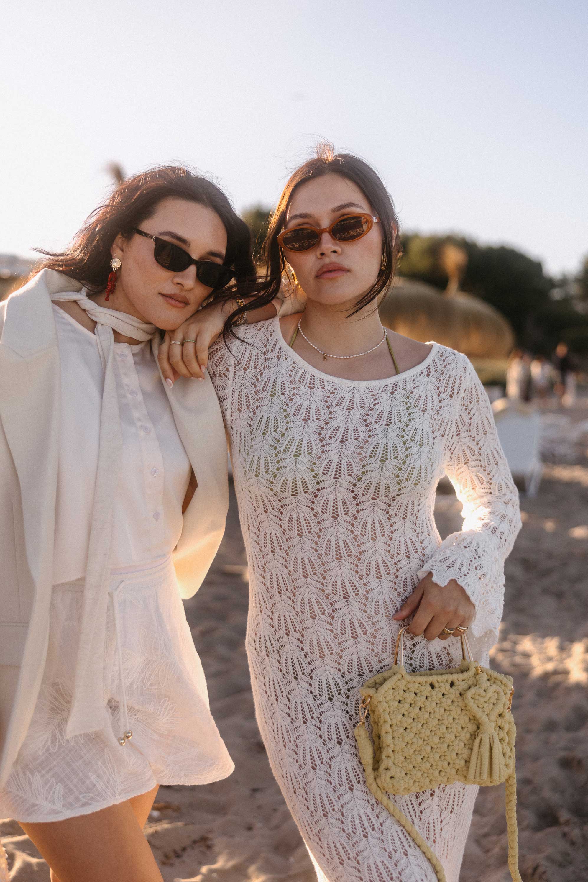 Zwei Frauen stehen bei goldenem Abendlicht am Strand und tragen sommerliche Looks in Cremeweiß. Die Frau rechts trägt ein langes Häkelkleid mit Glockenärmeln, Sonnenbrille und eine kleine geflochtene Tasche, während die Frau links einen hellen Blazer über einem weißen Outfit kombiniert. Der Sandstrand und die warme Abendsonne schaffen eine elegante, mediterrane Atmosphäre.
