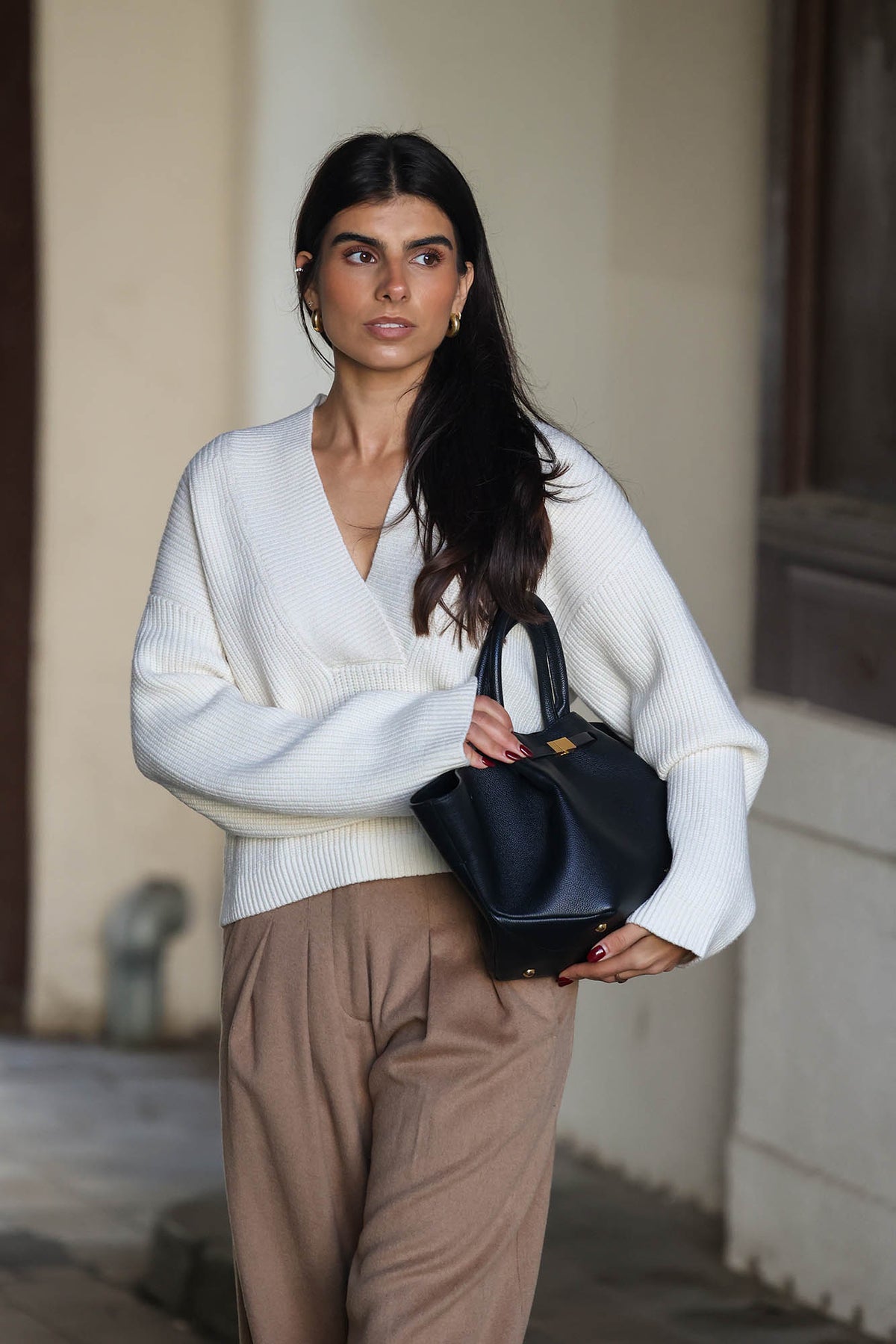 Frau trägt einen cremeweißen Strickpullover mit V-Ausschnitt und weiten Ärmeln, kombiniert mit einer braunen Hose und schwarzer Handtasche.