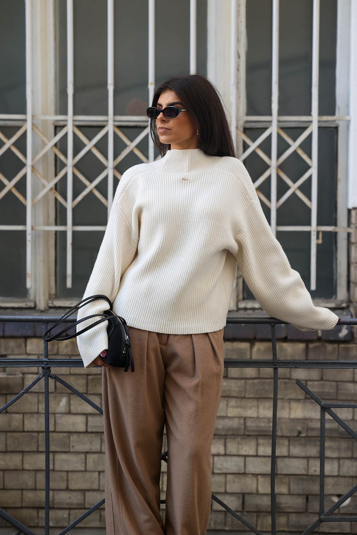 Frau trägt einen cremeweißen Strickpullover mit Stehkragen und weiten Ärmeln zu einer braunen Wide-Leg-Hose und schwarzer Handtasche.