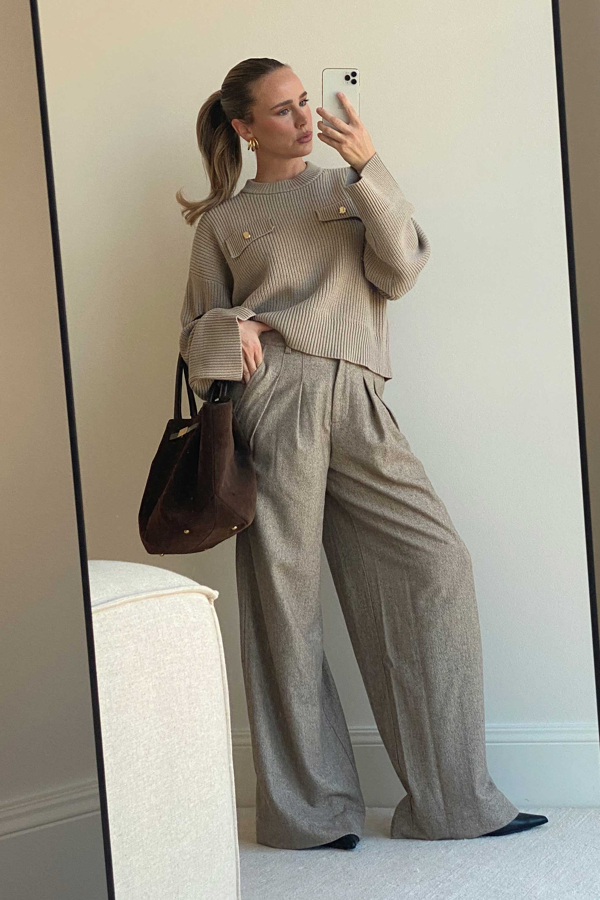 Frau macht ein Spiegelselfie in einem minimalistischen Raum. Sie trägt ein beigefarbenes, geripptes Strickoberteil mit aufgesetzten Taschenklappen und goldenen Knöpfen sowie eine weit geschnittene, graubraune Bundfaltenhose. In der Hand hält sie eine dunkelbraune Tasche aus Wildleder. Ihr Haar ist zu einem hohen Pferdeschwanz gebunden, sie trägt goldene Creolen.