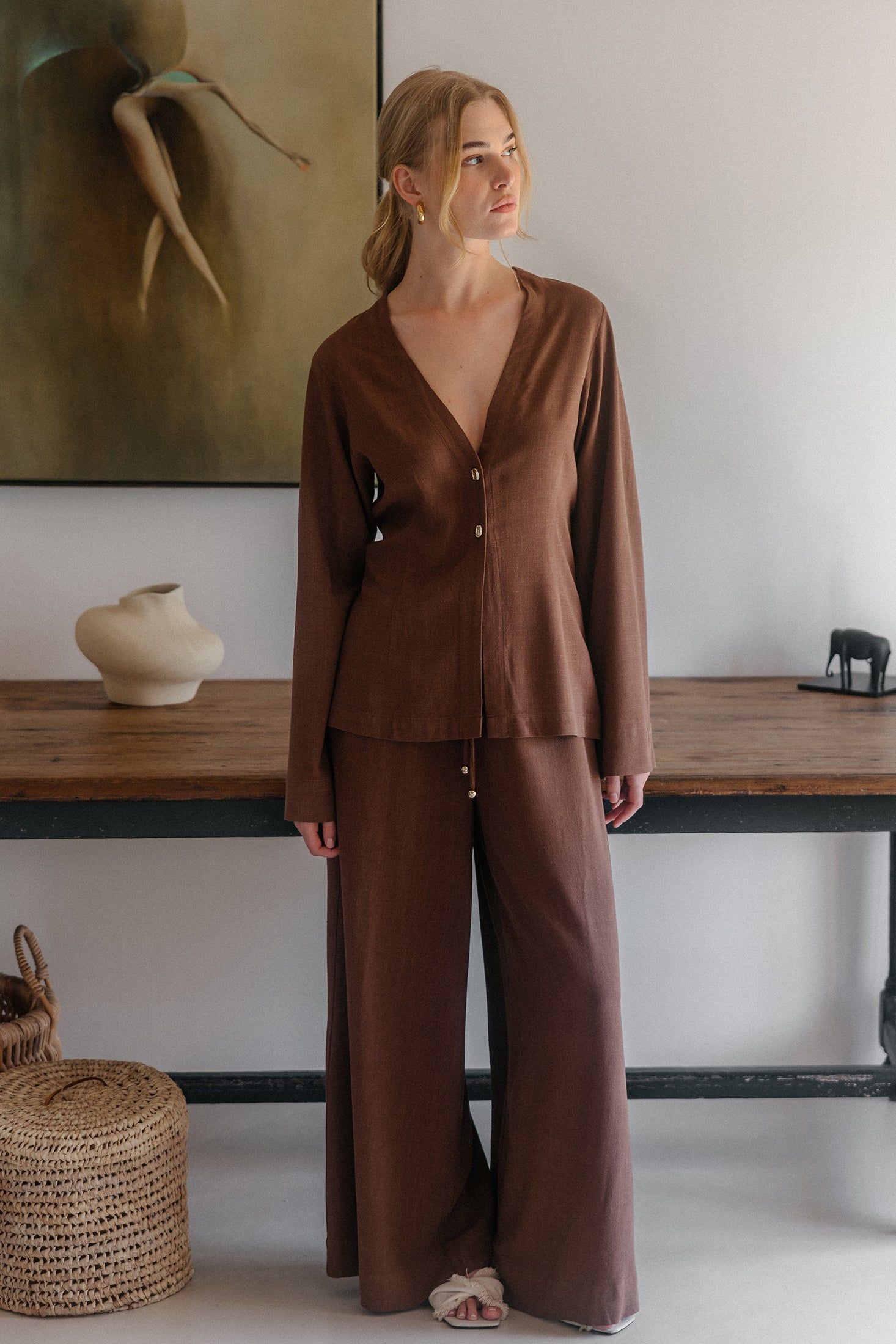 Model in braunem Zweiteiler mit tief ausgeschnittener Bluse und weiter Hose vor minimalistischer Inneneinrichtung mit Holztisch und Wandkunst.