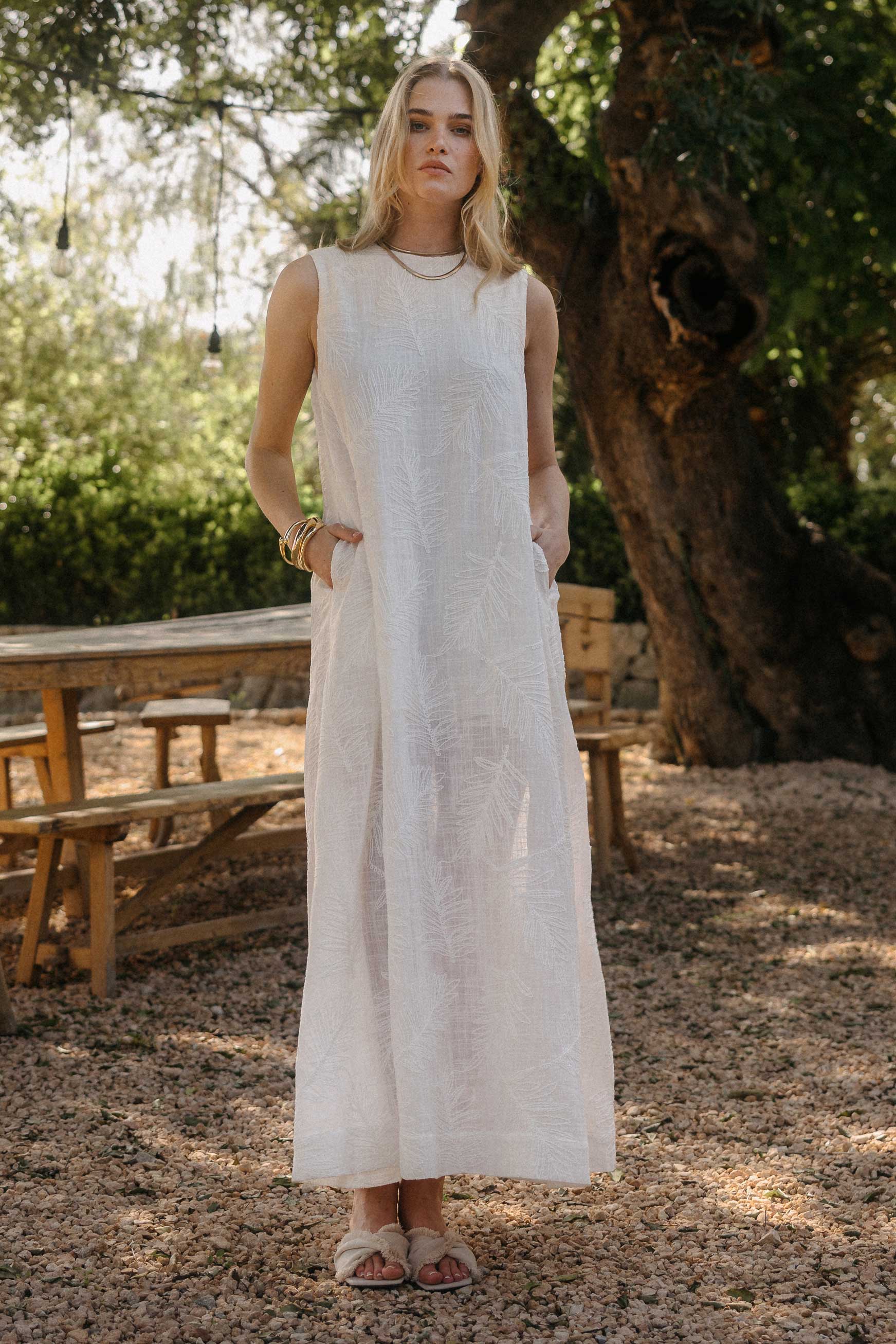 Model steht im Freien unter einem großen Baum und trägt ein langes, ärmelloses weißes Kleid mit geradem Schnitt und seitlichen Taschen, kombiniert mit Sandalen und goldenen Armreifen
