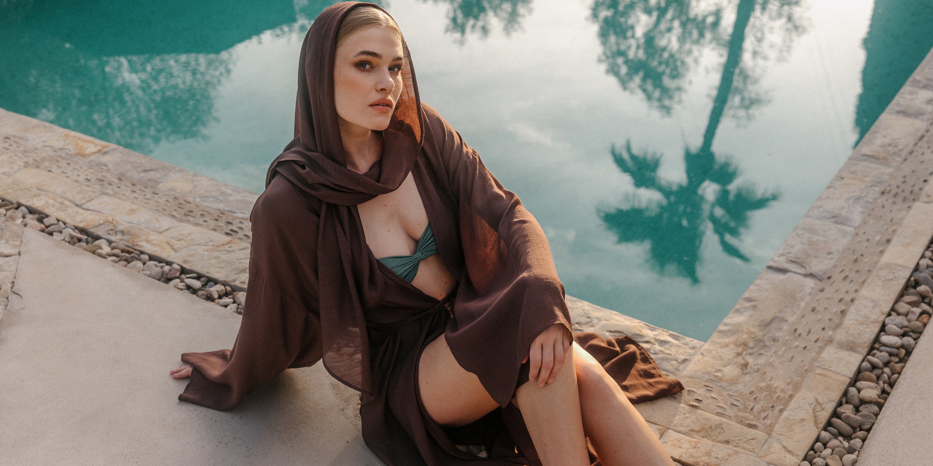 Model sitzt am Poolrand in einem transparenten dunkelbraunen Cover-up mit Kapuze über einem olivgrünen Bikini bei warmem Abendlicht