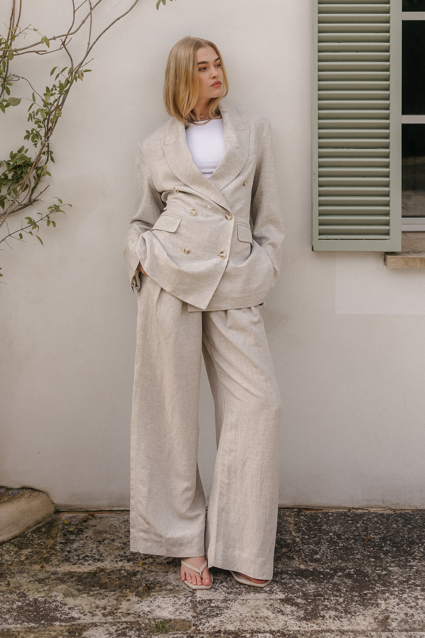 Model trägt ein zweiteiliges beigefarbenes Leinen-Set aus tailliertem doppelreihigem Blazer mit breitem Revers und weiter Hose, kombiniert mit weißem Top, goldfarbenem Halsschmuck und Sandalen