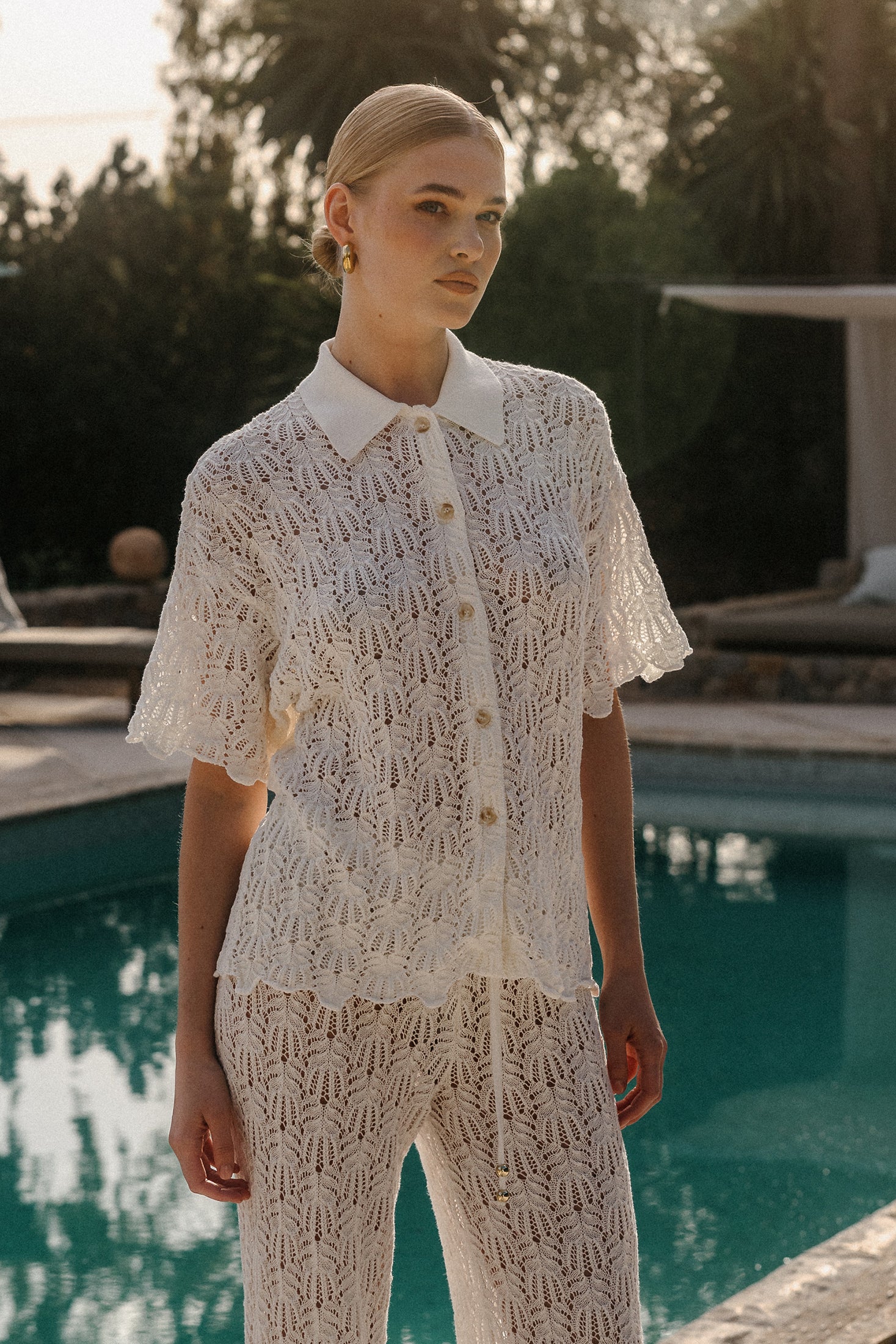 Model in weißem Häkel-Zweiteiler mit kurzärmeliger Knopfbluse und weiter Hose am Pool bei warmem Sonnenlicht.